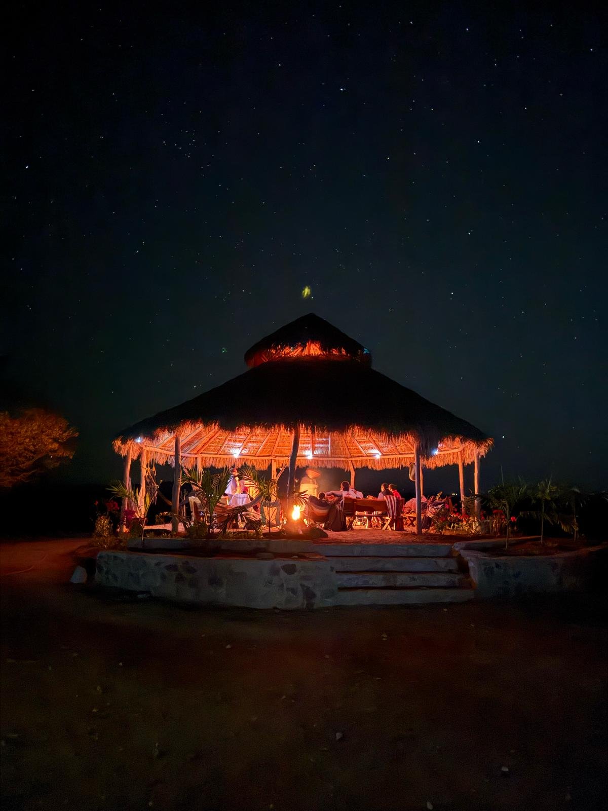 Nighttime scene of an open-air hut with thatched roof, lit by a fire, and people sitting at tables outside, under a starry sky.