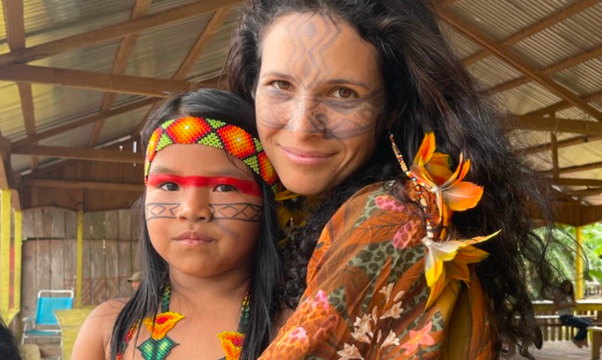 A woman and a young girl dressed in traditional indigenous clothing, with face paint, jewelry, and floral accessories, standing inside a wooden structure.