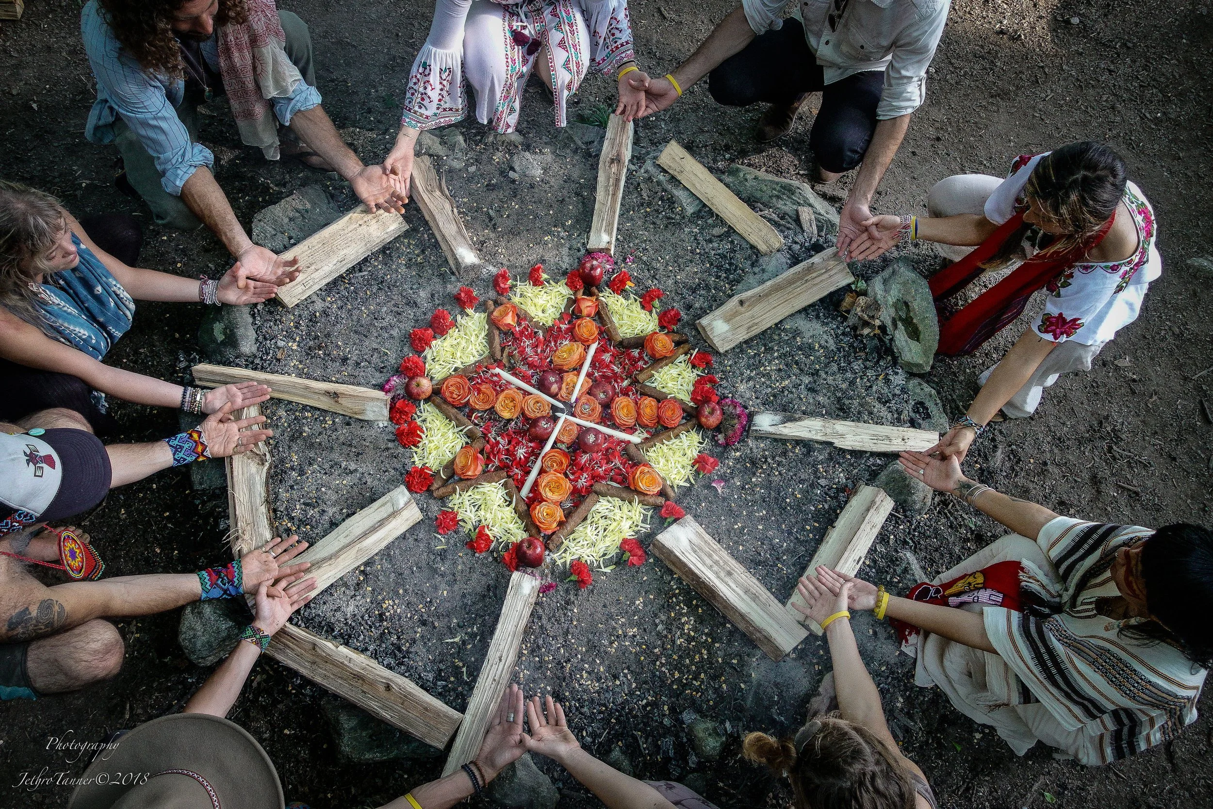 A group of people holding hands in a circle around a colorful floral arrangement that resembles a star, placed on the ground with wooden sticks forming the star's outline.