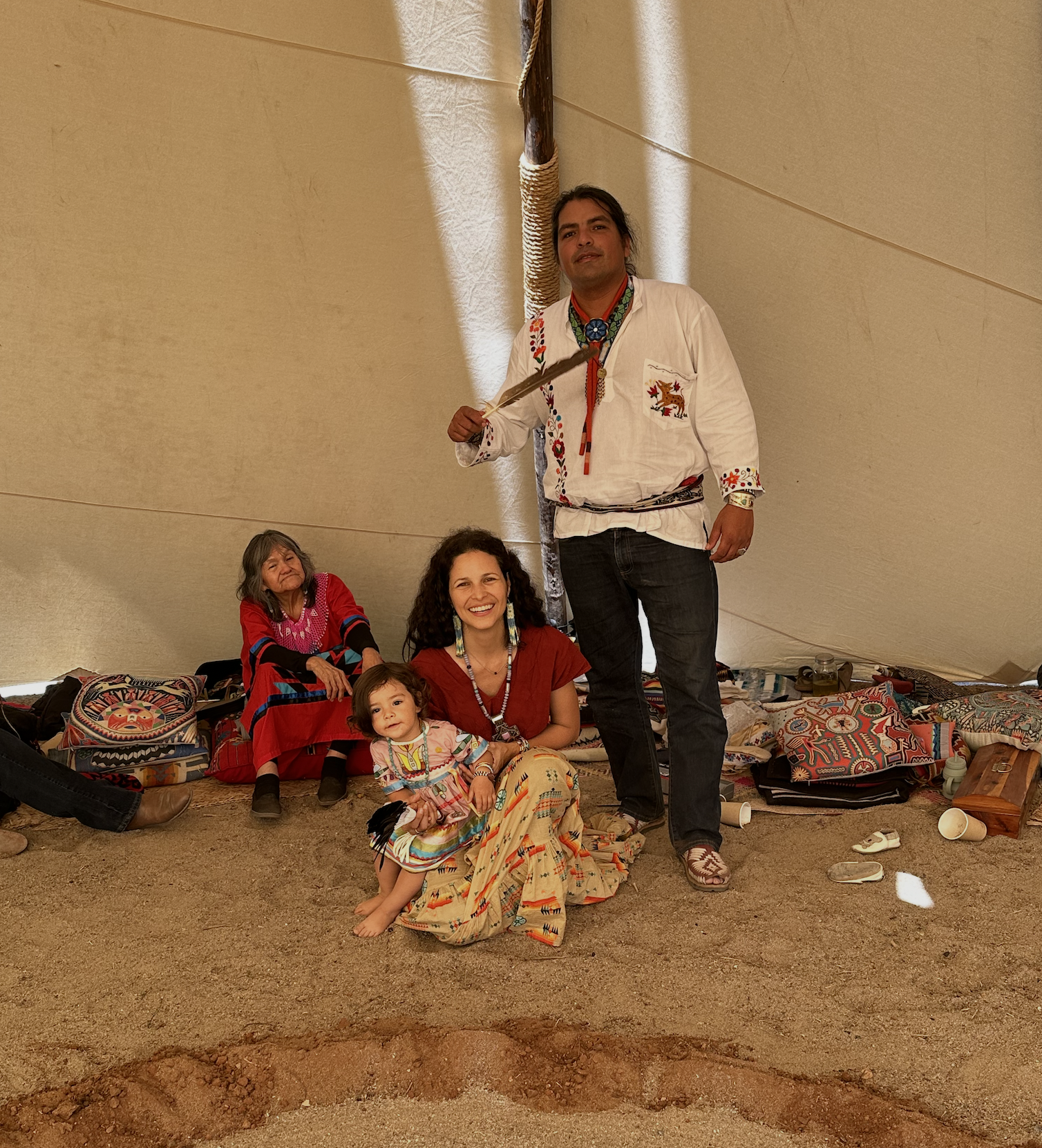 A group of four Native American individuals inside a large beige tent, with two women sitting on the ground and two men standing, wearing traditional clothing and accessories, surrounded by patterned bags and cups.