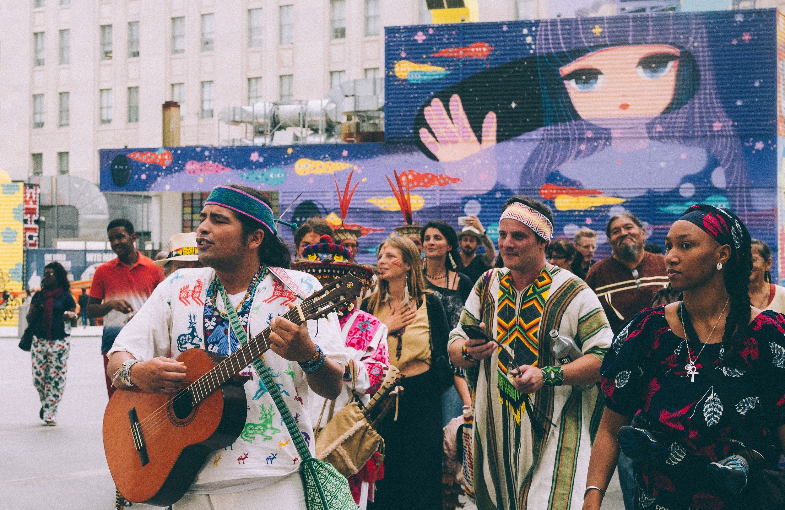 Group of people at a cultural event in an urban setting, some wearing traditional attire, with a large mural of a girl with purple hair and colorful fish in the background, and a musician playing guitar.