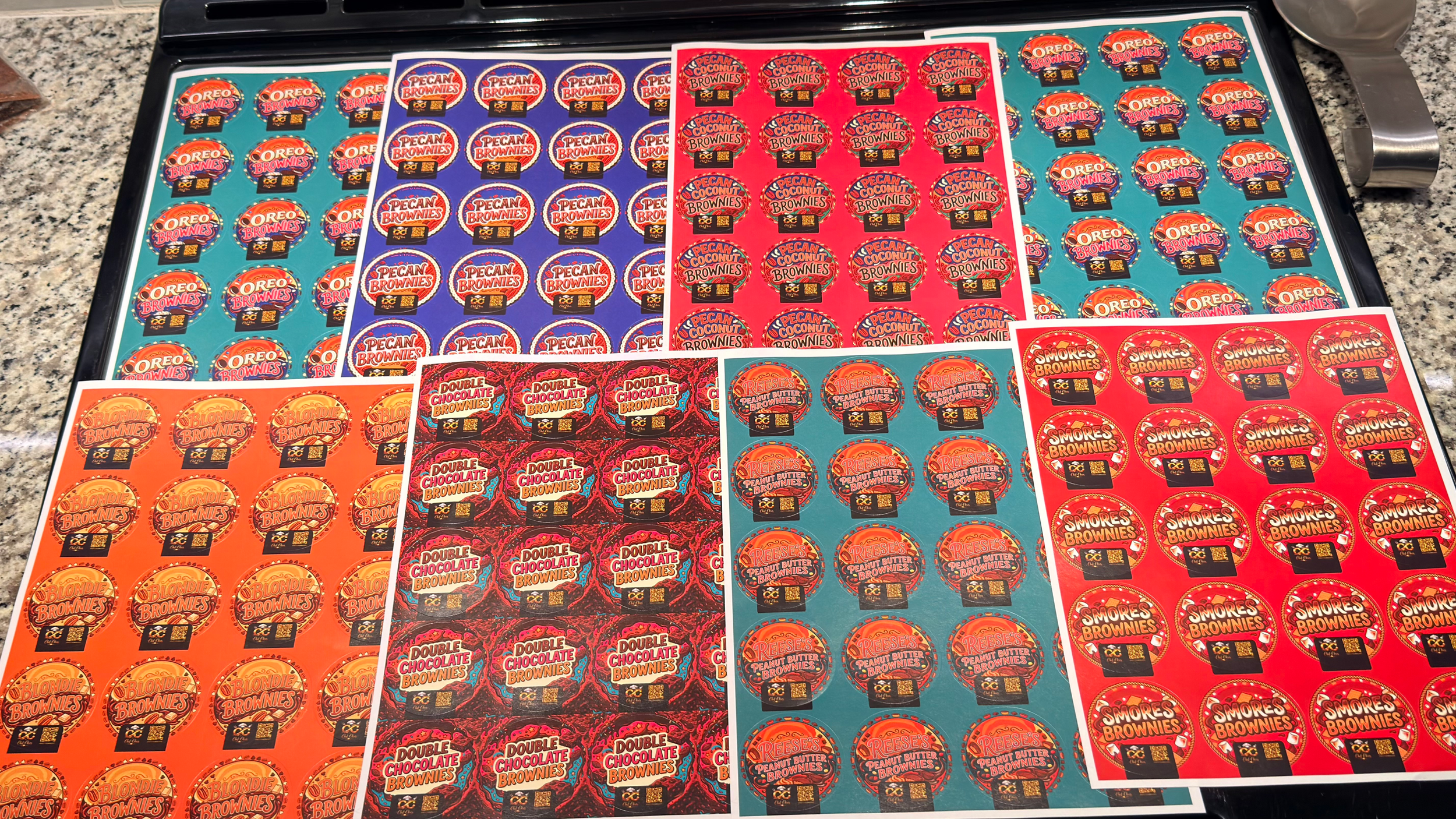 Six sheets of stickers featuring various ice cream flavors, including Oreo Brownies, Pecan Brownies, Blondie Brownies, Reese's Peanut Butter, and S'mores Brownies, arranged on a black tray on a granite countertop.
