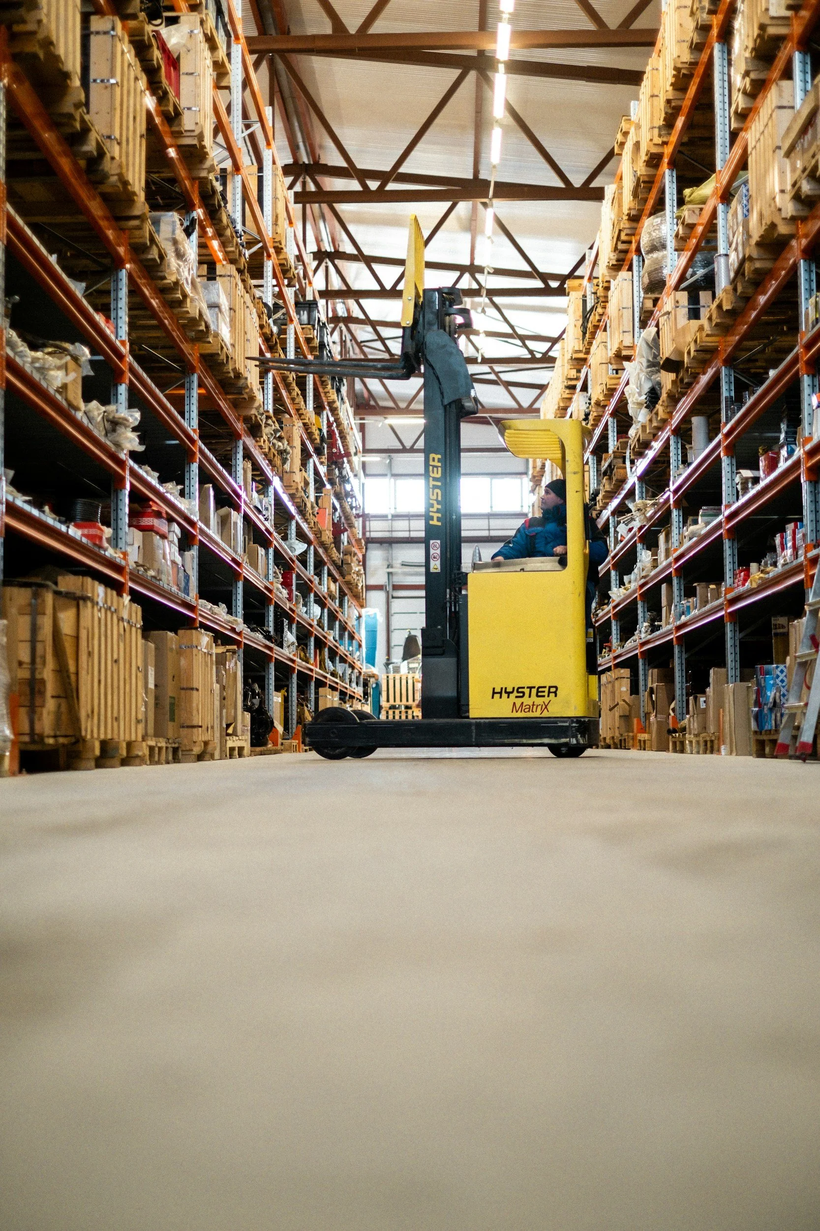 A man operating a yellow Hyster forklift in a warehouse aisle with tall shelves filled with various boxes and items.