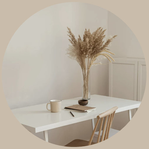 A white table with a beige mug, a notebook, a pen, and a tall glass vase with dried pampas grass in a minimalist room.
