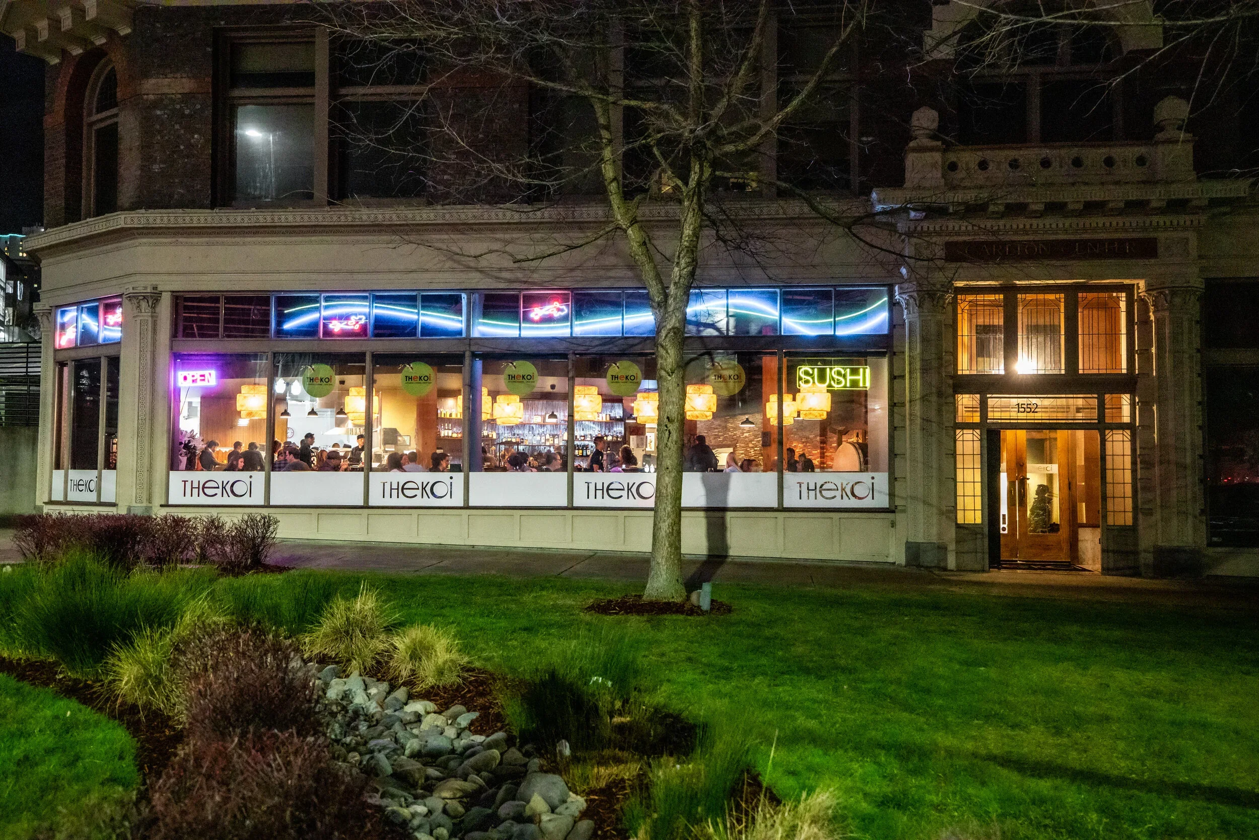 A restaurant with large glass windows showing the interior with tables and hanging lamps at night. Neon signs advertising sushi and other drinks. A tree in the foreground with a small landscaped area and grass.