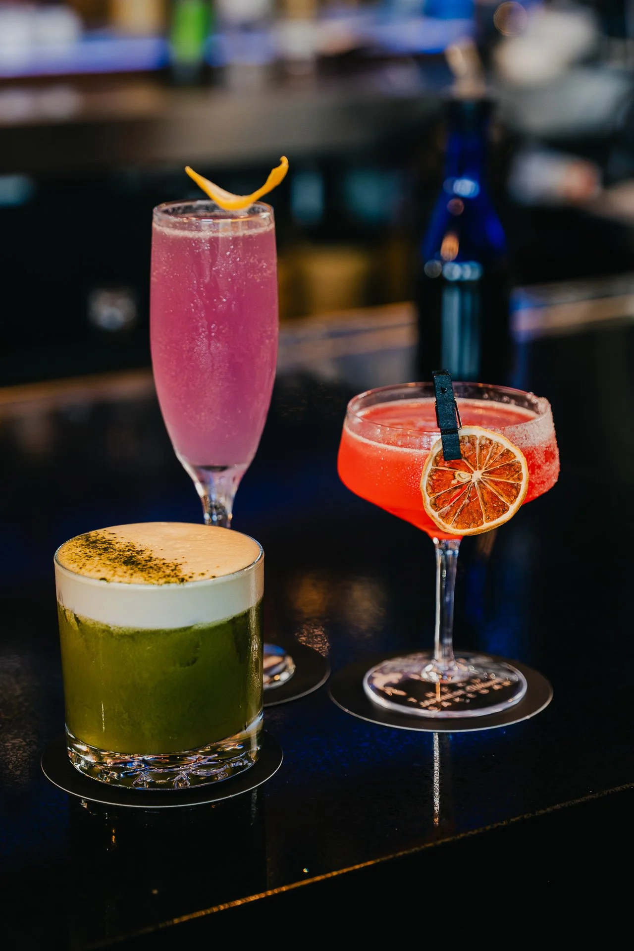 Three colorful cocktails on a bar counter: a green drink with foam and sprinkled herbs at the front, a pink bubbly drink in a tall flute glass at the back, and a red cocktail garnished with a lemon slice and black clip on the right.