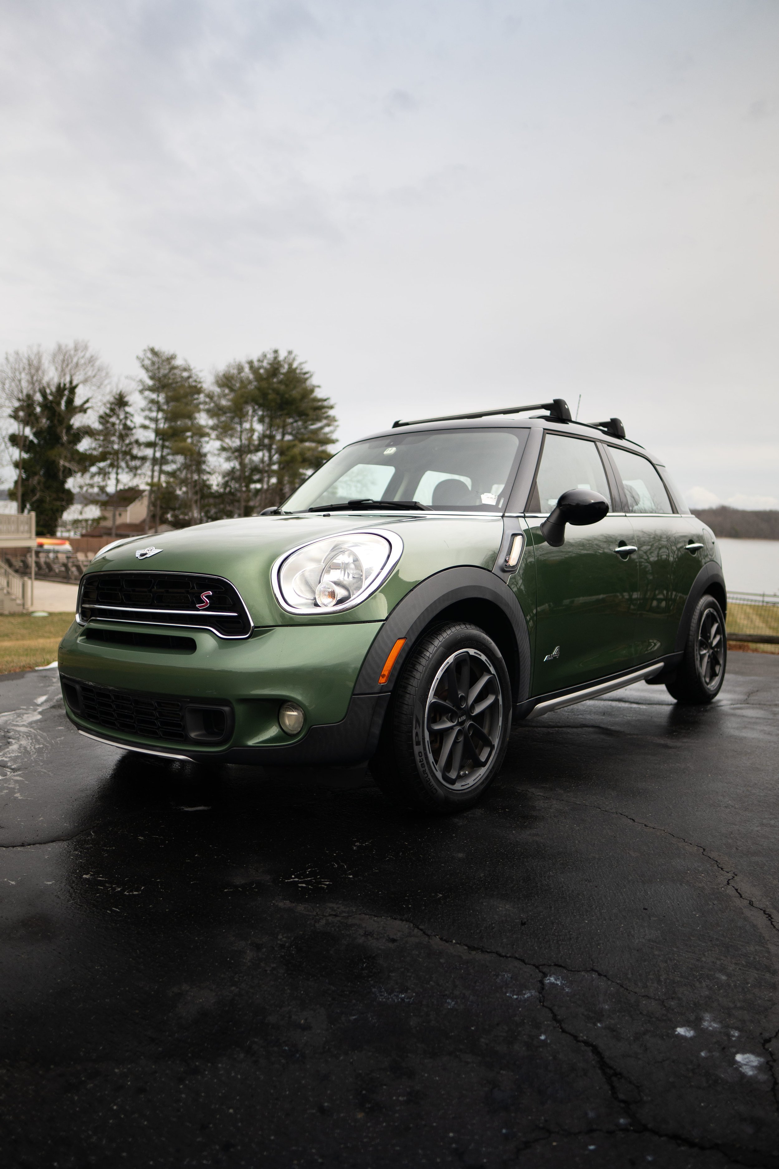 A green Mini Cooper car parked on a black asphalt surface near a body of water with trees and houses in the background under a cloudy sky.