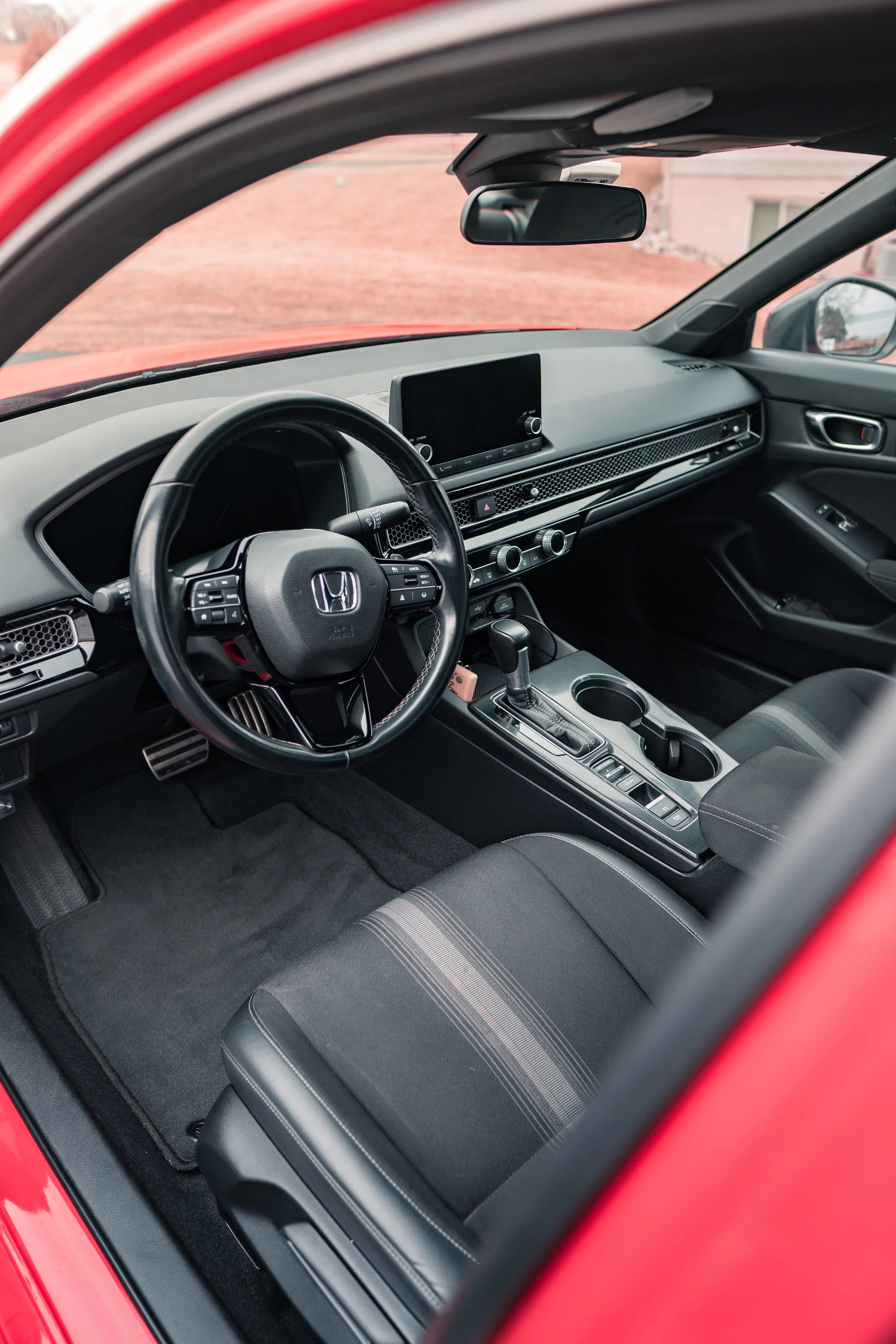 The interior of a red Honda car showing the dashboard, steering wheel, center console with gear shift, and front seat with fabric upholstery.