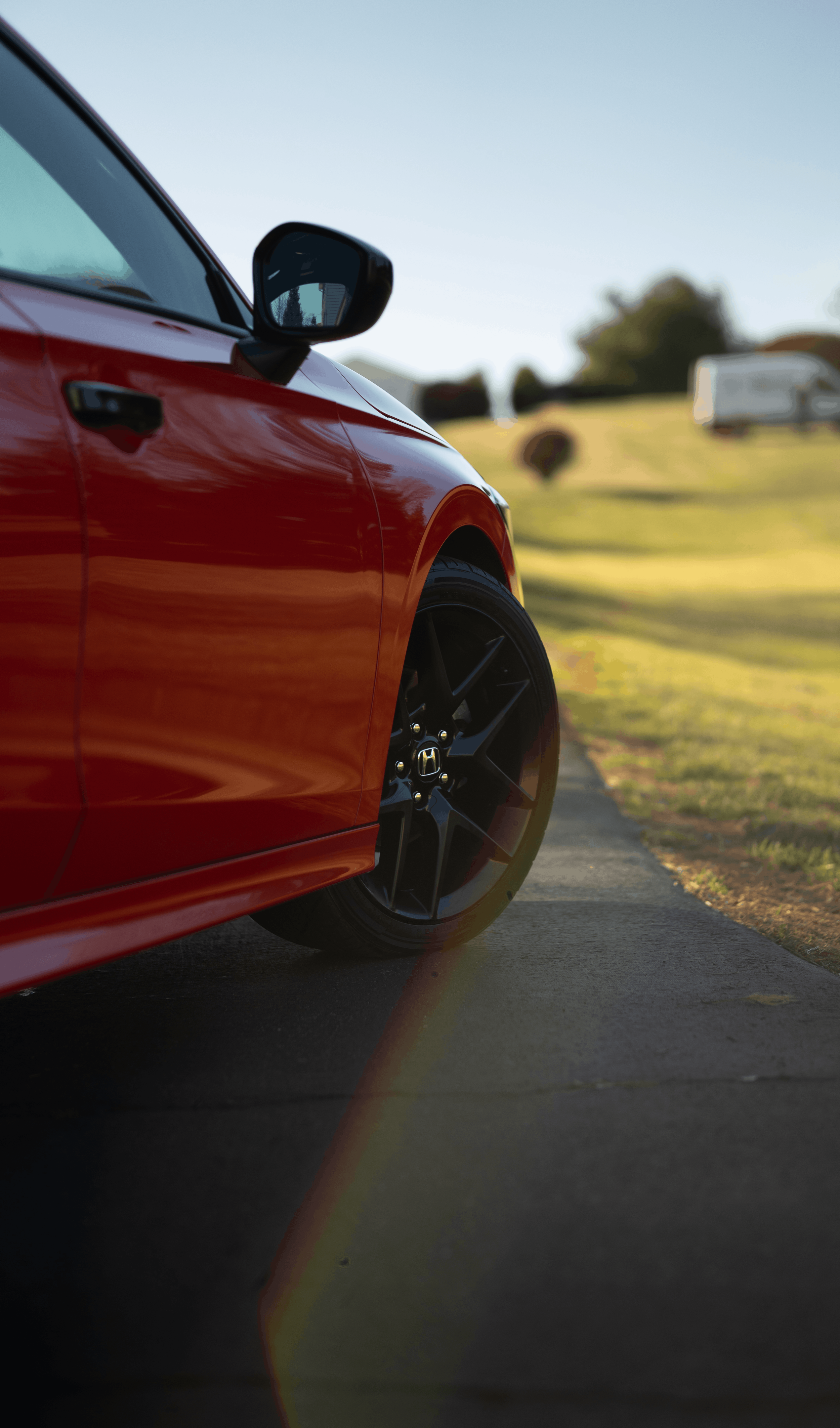 Close-up of the front side of a red Honda car parked on a driveway with a background of grassy hill and a white trailer.