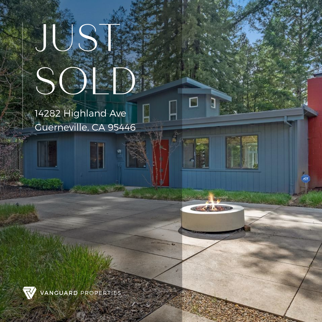 A modern blue house with a red door and a small tower structure in a wooded area. The front yard has a concrete patio with a fire pit and surrounding greenery. The photo indicates the house has just been sold and displays the address 14282 Highland A