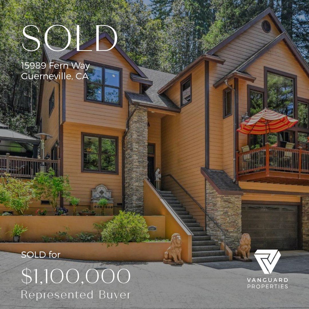 Front view of a modern, multi-story house with tan siding and stone accents. The house features large windows, a staircase leading to the entrance, and a balcony with a red-and-orange striped umbrella. There are two lion statues at the base of the st