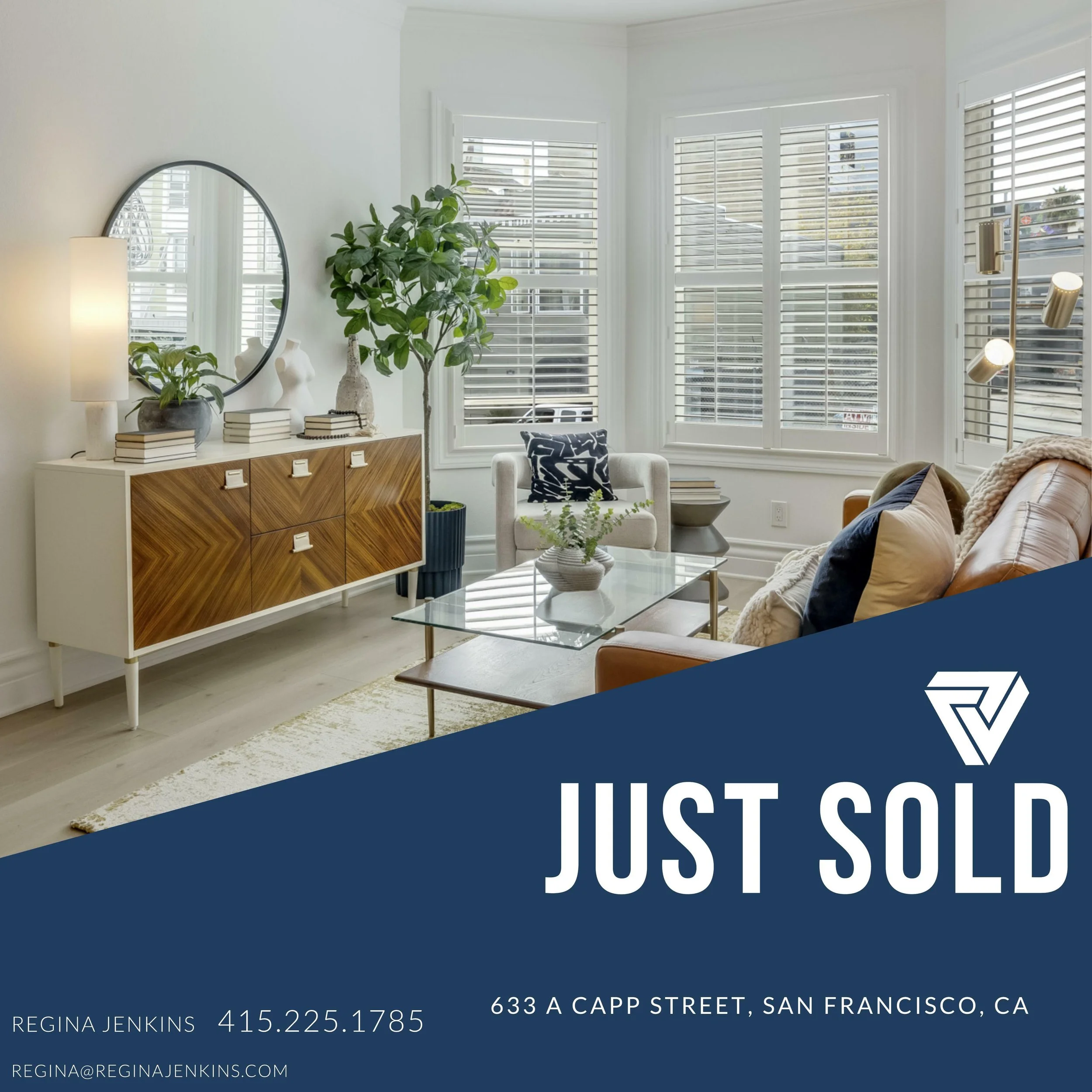 Interior of a living room with white walls, large bay windows with blinds, a white armchair with a black and white pillow, a tan leather sofa with pillows, a glass coffee table, a wooden sideboard with plants and decorative objects, a round mirror, a