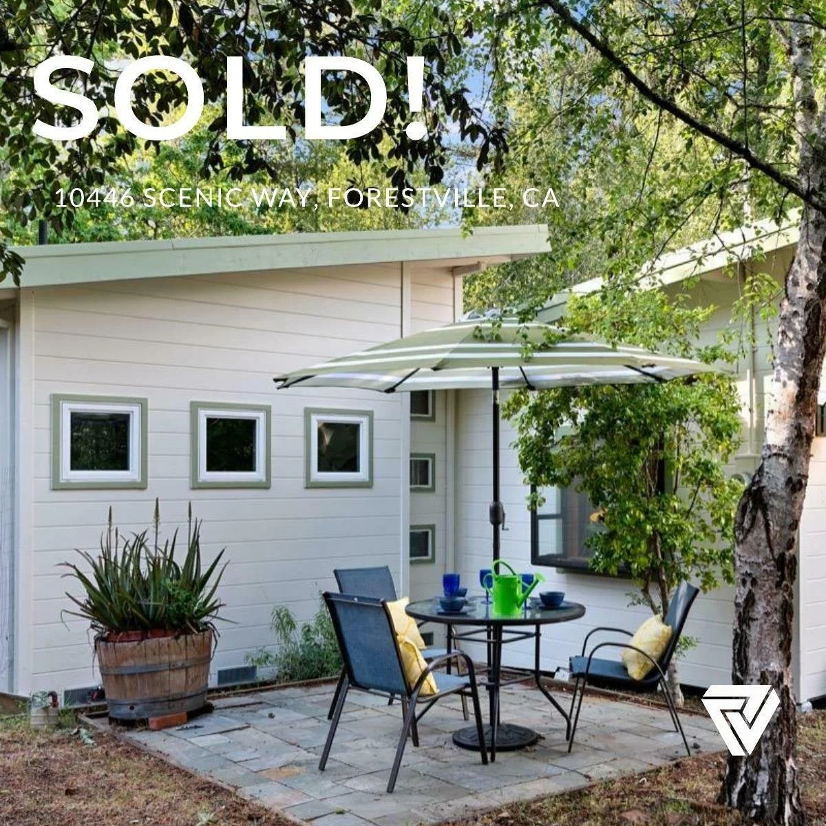 A backyard patio with a round table, four chairs with cushions, a large umbrella, potted plants, and a white house with multiple small windows. The setting looks peaceful and inviting with trees providing shade.