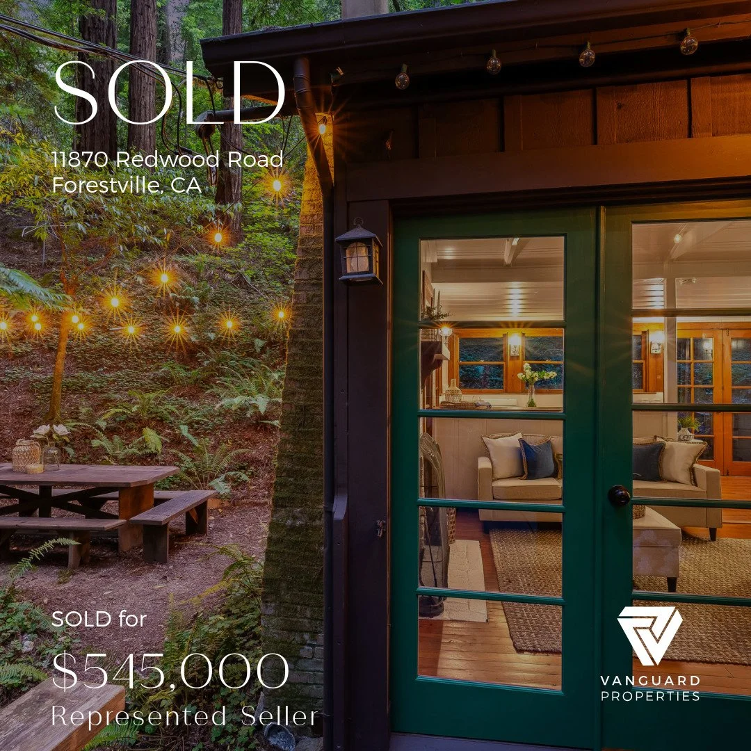 Interior view of a cozy living room seen through a glass door at sunset, with a forested outdoor area decorated with string lights and a picnic table.