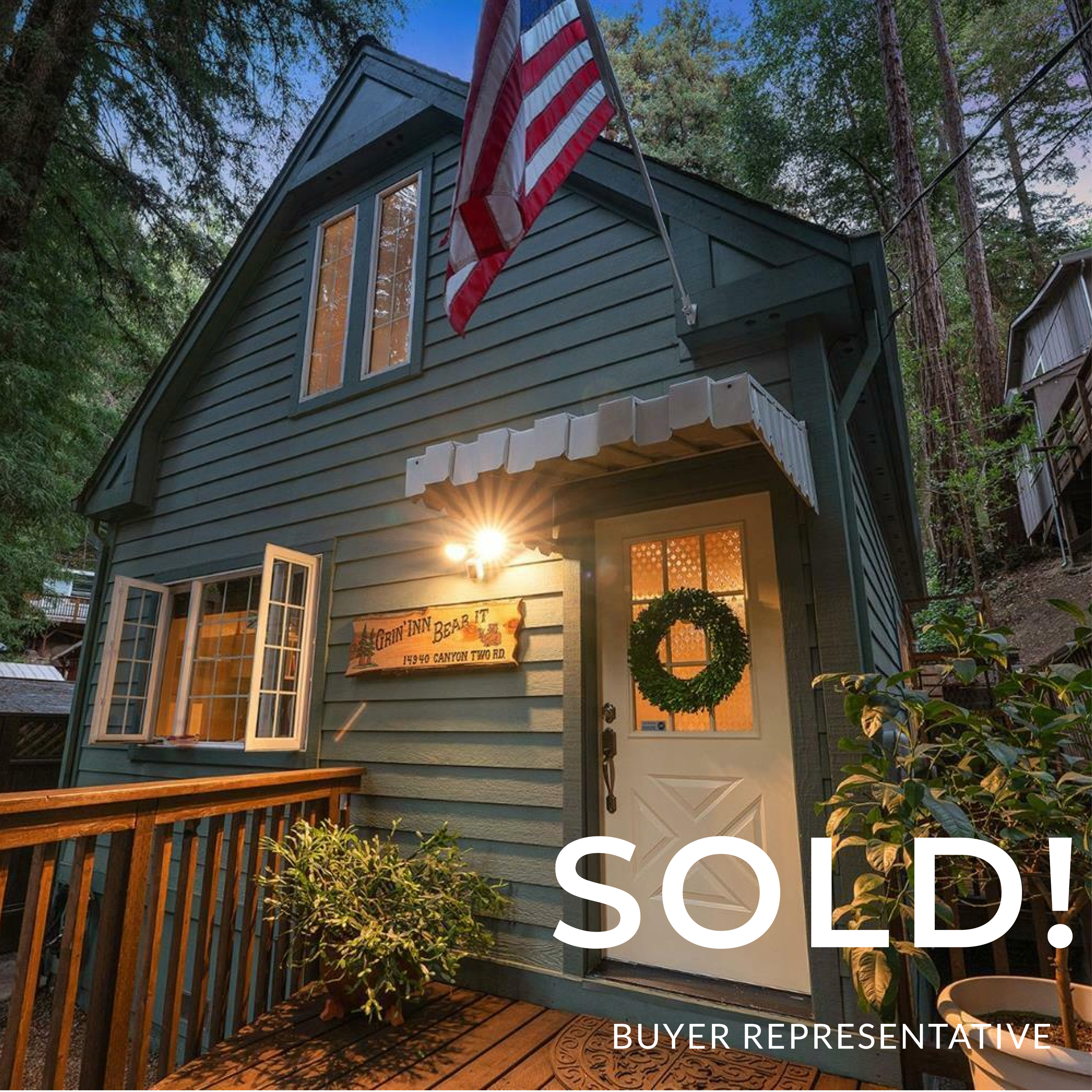 A two-story house painted blue with a wooden deck and a green wreath on the front door, decorated for the holidays. An American flag is mounted on the house, and a sign reading 'The Grin Inn Bear It' with an address 15940 Canyon Two Rd is displayed n