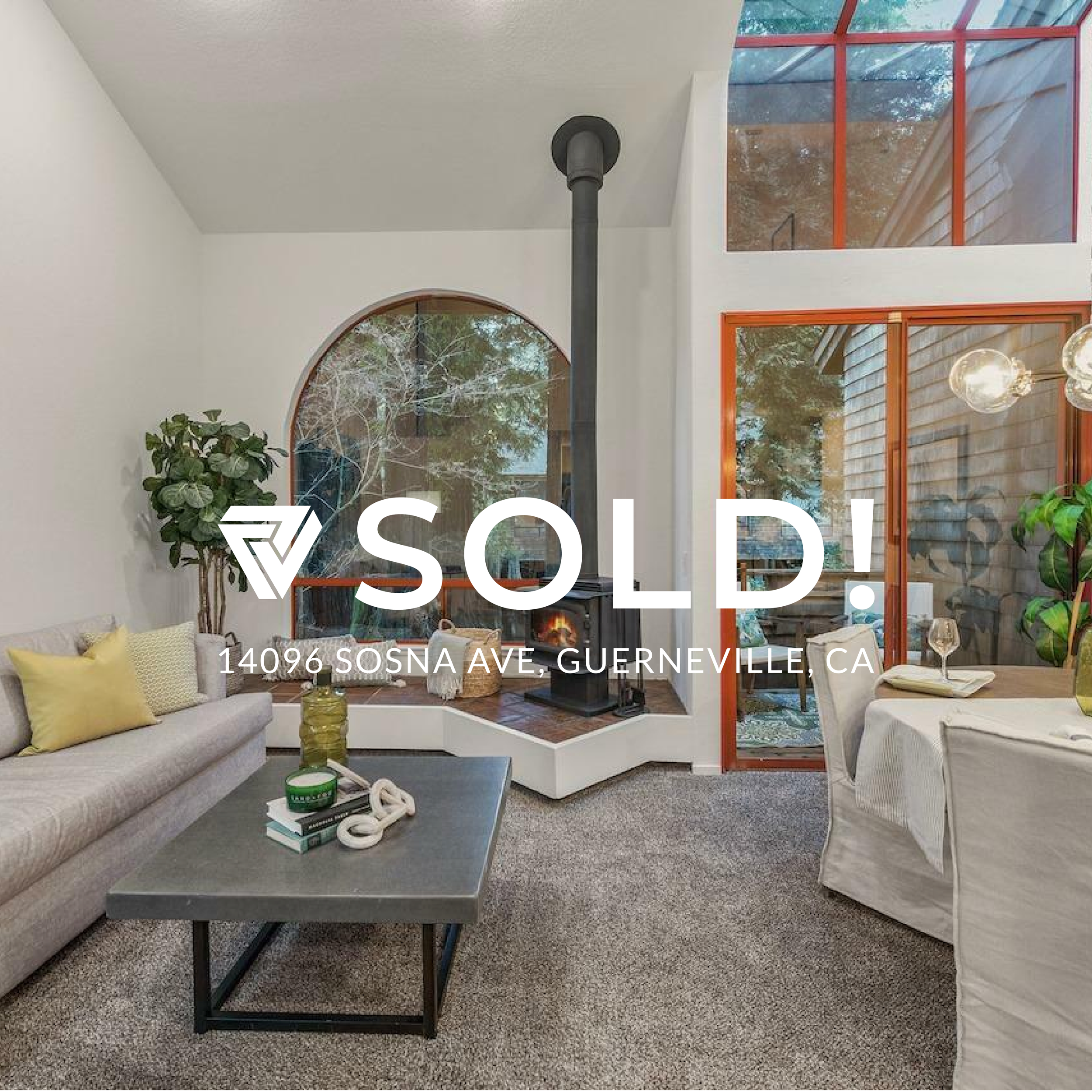 Living room with beige sofa with yellow and green pillows, black coffee table with books and decorative objects, wood stove with pipe, large arched window, and sliding glass door with orange trim leading to outdoor area with wooden walls and trees.