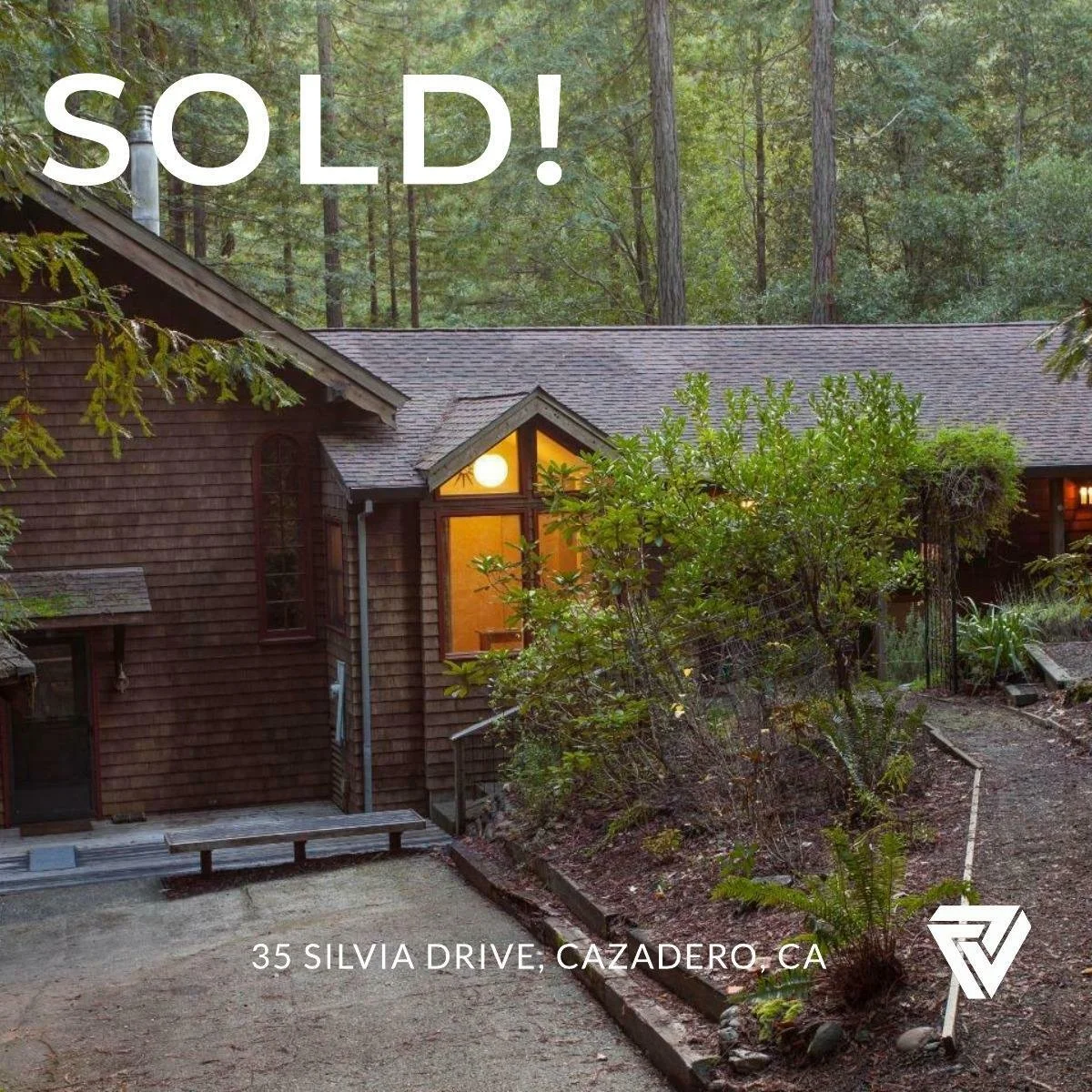 A house in the woods with warm lights on, a gravel driveway, surrounding trees, and a sign on the ground that reads 'sold!' with the address 35 Silvia Drive, Cazadero, CA.