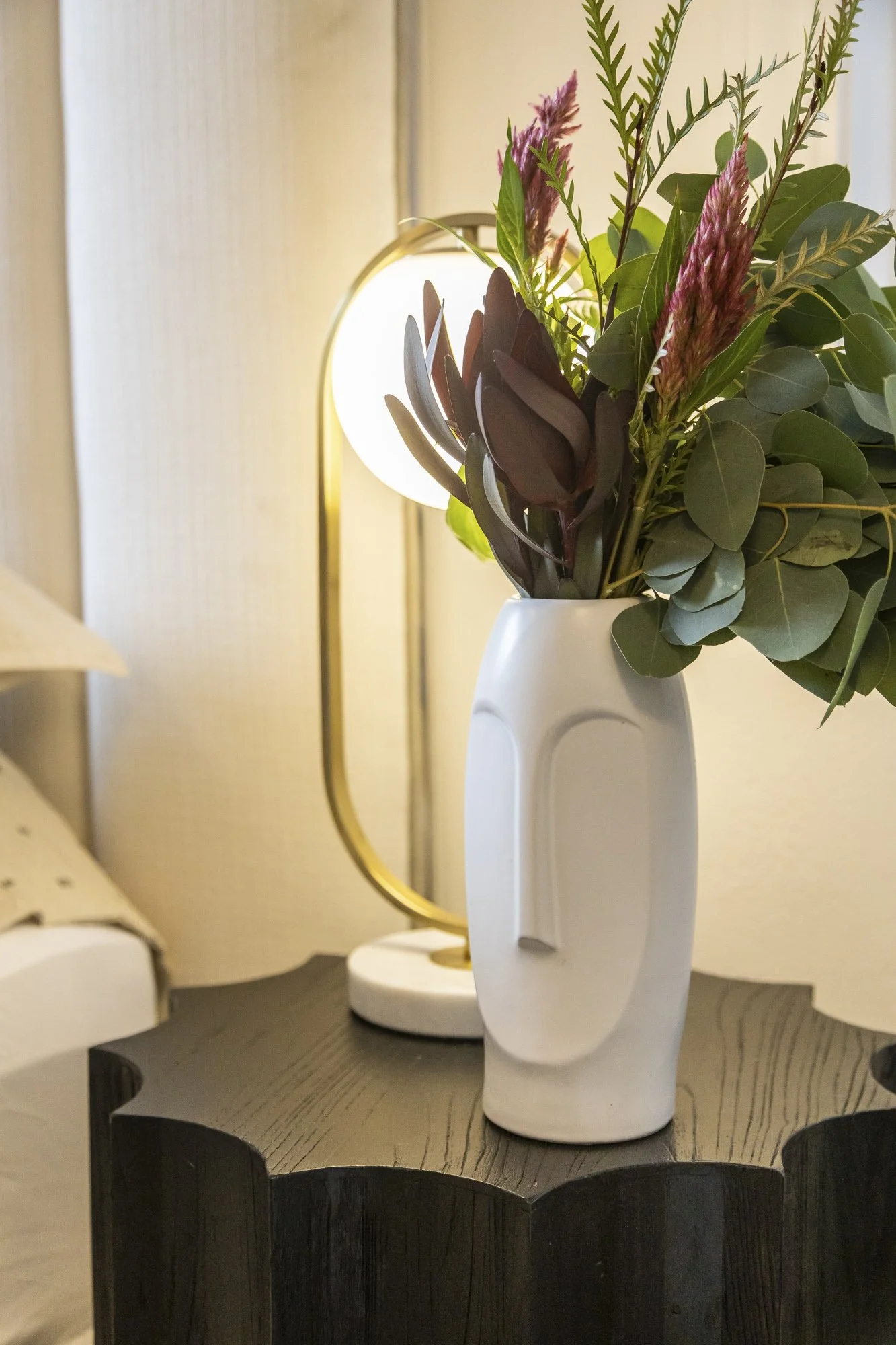 Close-up of a modern white ceramic vase with a face design, holding a bouquet of green leaves and pink flowers on a black wooden table, with a brass table lamp and beige wall in the background.