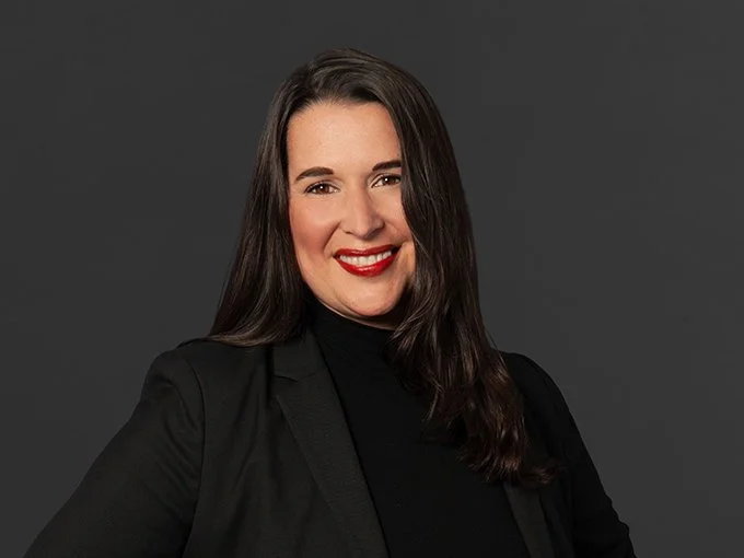 A woman with dark brown hair, red lipstick, and a black blazer smiling against a gray background.