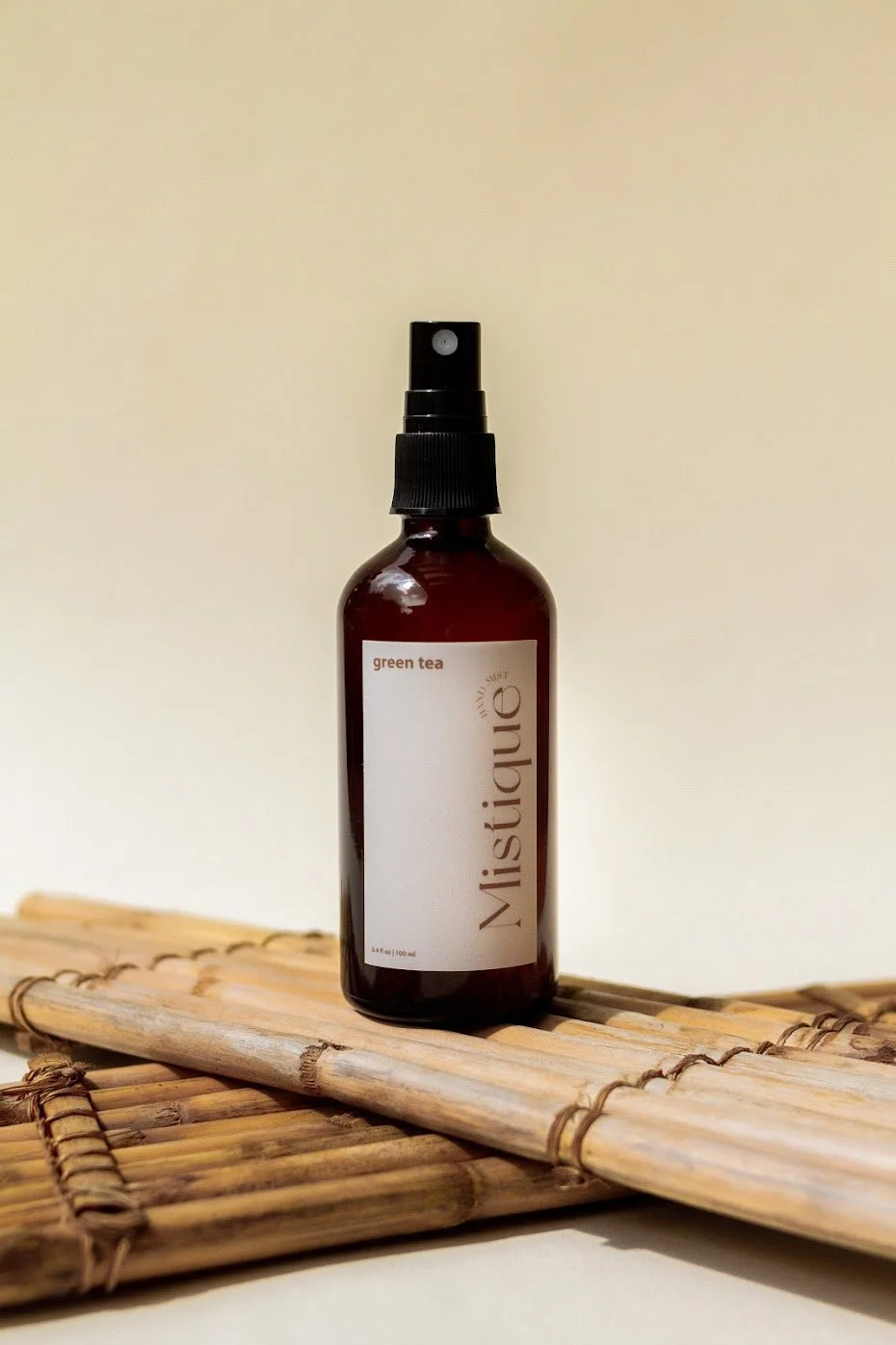 A brown spray bottle labeled "Mistique" with "green tea" and "3.4 fl oz (100 ml)" on a white background, placed on a bamboo mat.