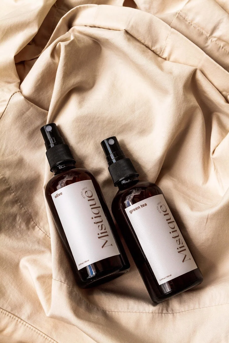 Two amber spray bottles labeled "Mistique," one with a white label reading "olive" and the other with a white label reading "green tea," resting on beige fabric.