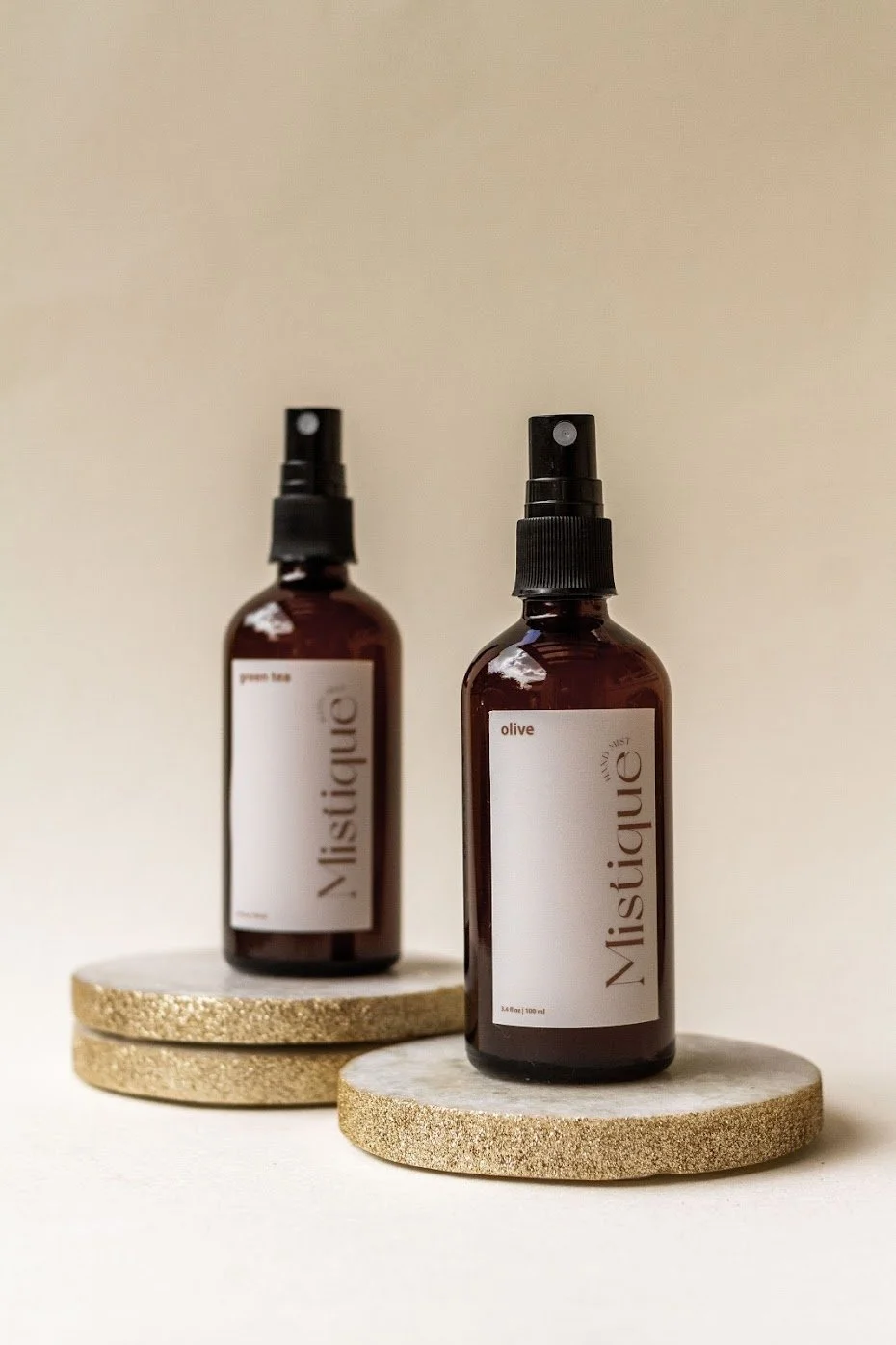 Two amber spray bottles labeled 'Mistique' with one labeled 'olive' and the other 'orange '., placed on round, textured coasters against a plain background.