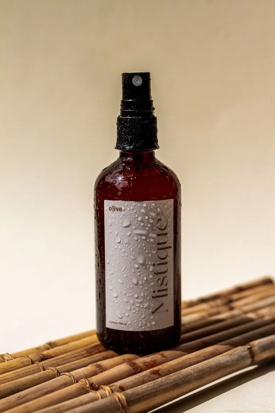A spray bottle labeled "Nistqure" with a white label and the word "olive" on it, sitting on a wooden mat, with water droplets on the bottle, against a plain background.