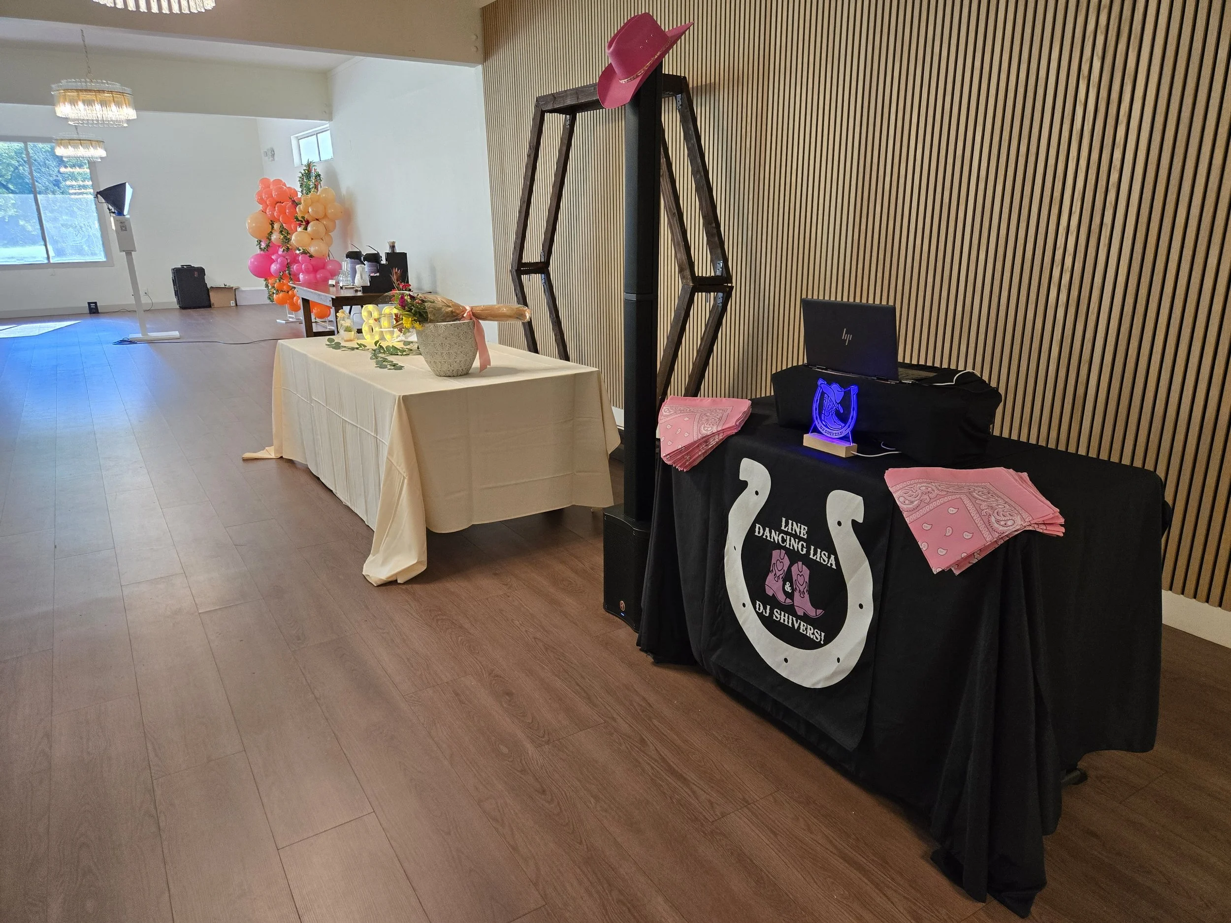 Private Event setup for a birthday party with a table of pink and orange balloons, a flower arrangement, and a music DJ station with pink bandanas, a laptop, and neon sign, inside a room with wooden wall panels and hardwood flooring.