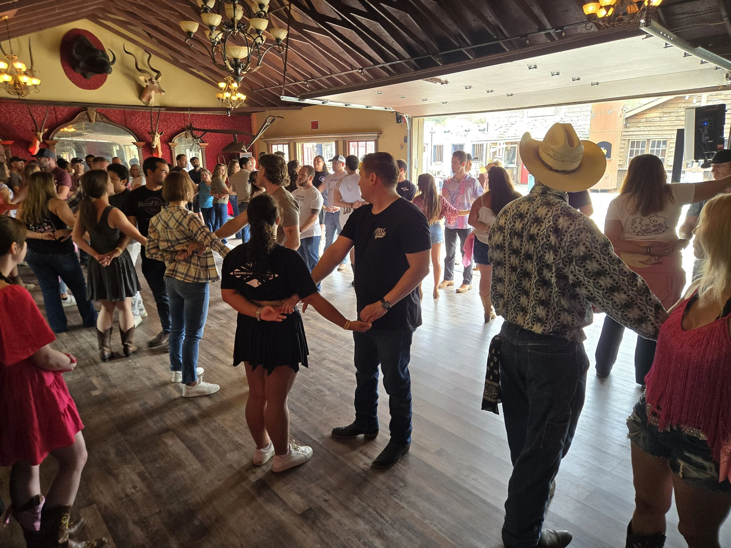 Country Dance Day in Half Moon Bay Event. Happens every August at Long Branch Saloon & Farms. Put on by Line Dancing Lisa. People dancing in a line inside a rustic-themed venue with wood floors, red wallpaper, and mounted animal heads on the walls.