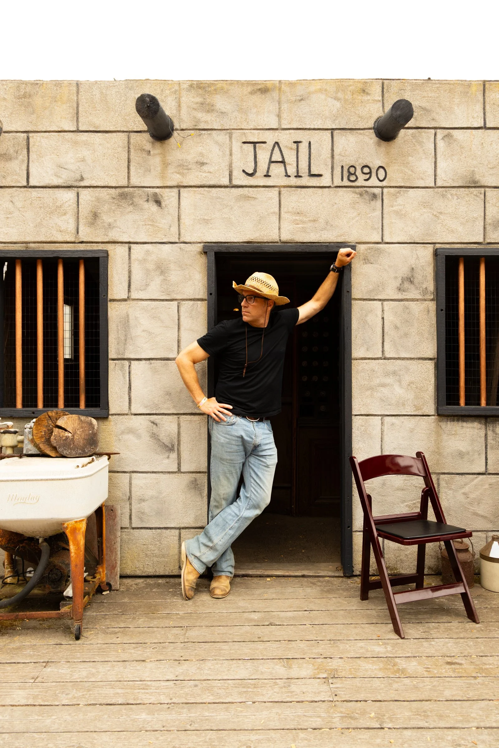Man in cowboy hat leaning against a jail door, with 'JAIL 1890' sign above, on a wooden porch with chairs and vintage objects. Country Dance Day in Half Moon Bay Event. Happens every August at Long Branch Saloon & Farms. Featuring DJ Shivers.