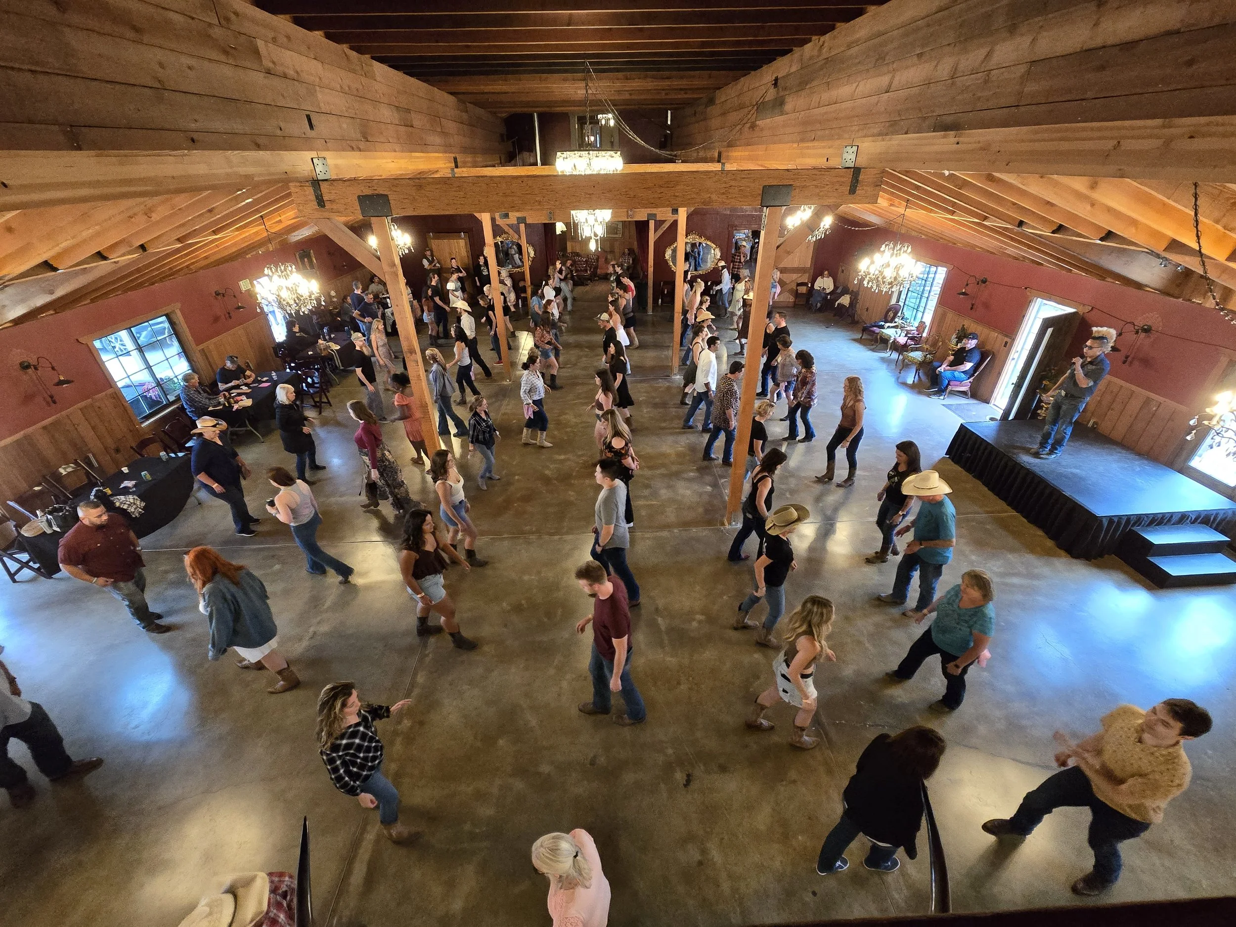 People line dancing in a rustic wooden hall with chandeliers and large windows, some wearing cowboy hats at Country Dance Day in Half Moon Bay featuring Line Dancing Lisa and DJ Shivers.