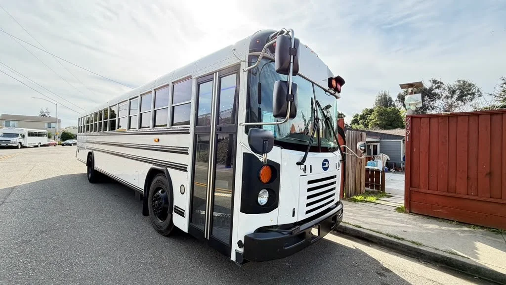 White school bus parked on the side of the road with a red wooden fence and some buildings in the background.