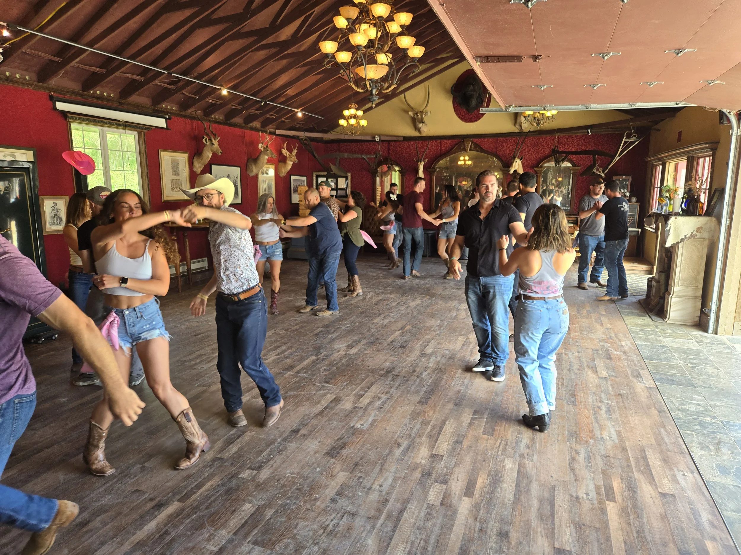 People dancing in a rustic, country-style indoor venue with red walls, mounted deer heads, and vintage decor. Country Dance Day in Half Moon Bay Event. Happens every August at Long Branch Saloon & Farms. Featuring all day line dancing lessons