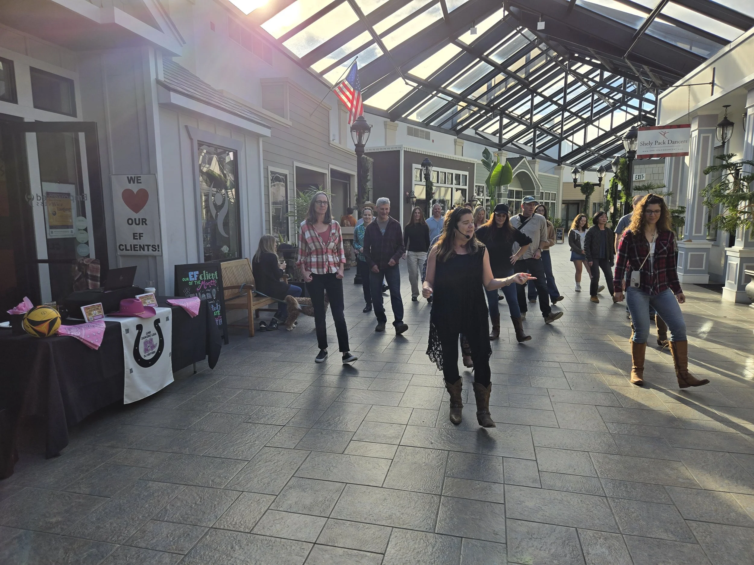 Private surprise birthday party with Line Dancing Lisa. People participating in a line dance inside a shopping mall with glass ceiling, some tables and signs on the left, and an American flag hanging from the ceiling.