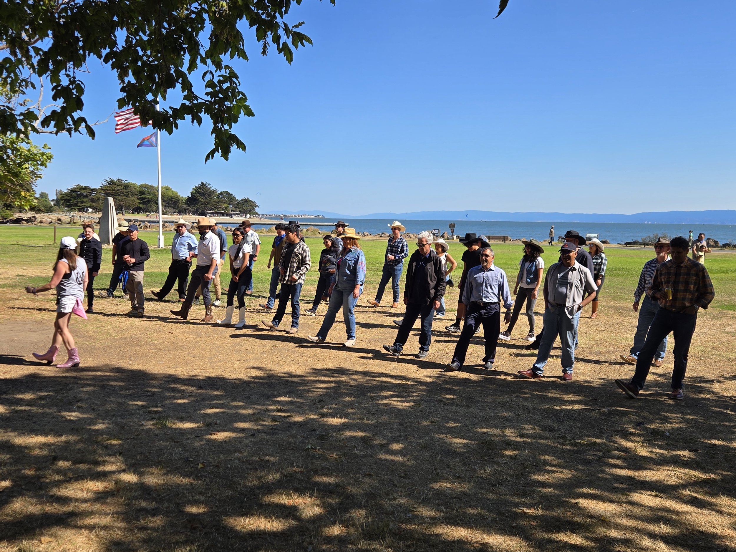 Line Dancing Lisa and DJ Shivers outdoor Corporate retreat event. Group of people walking on a grassy area near the waterfront, with American and state flags on flagpoles, trees, and water in the background, on a sunny day.