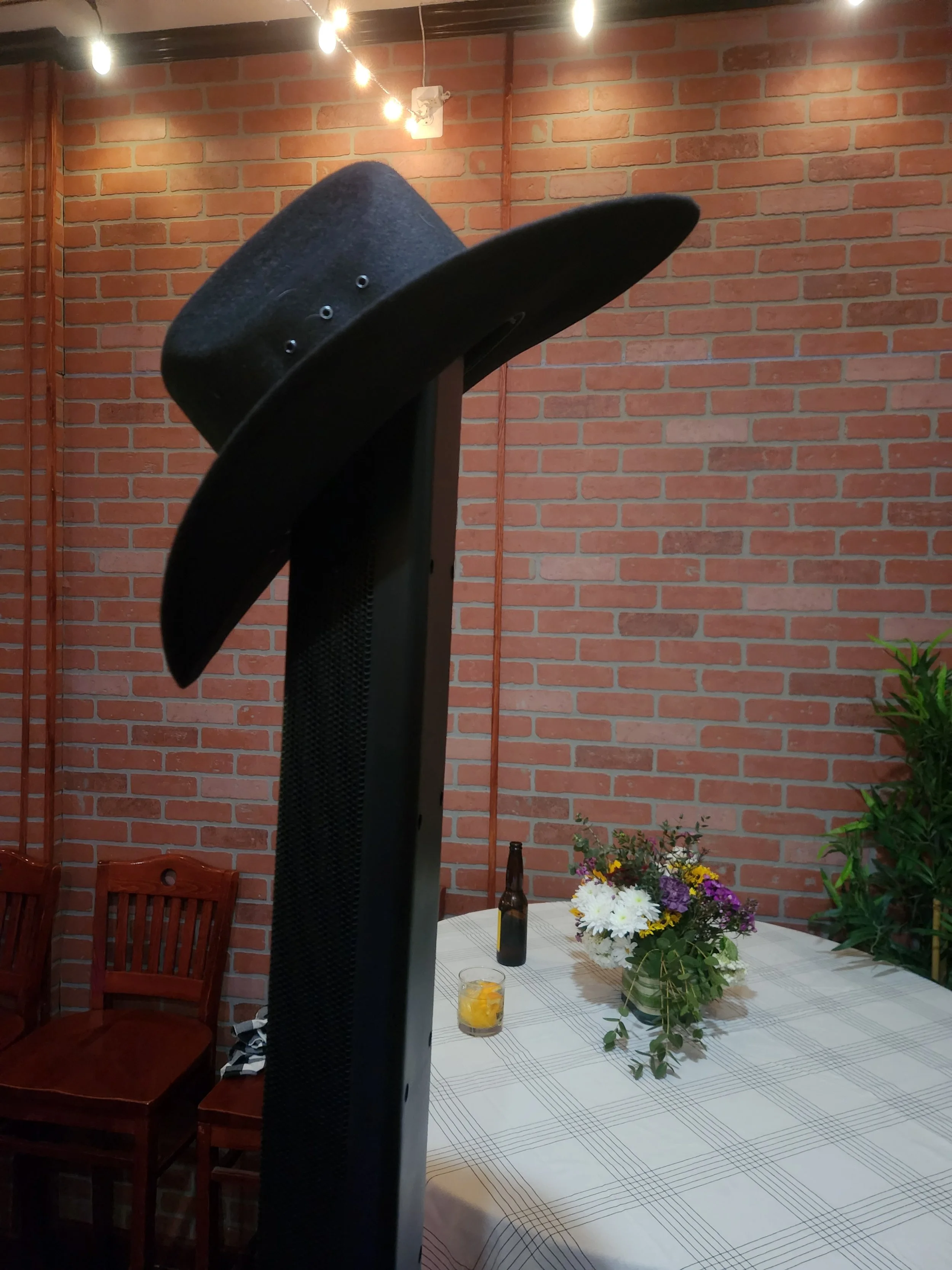 A photo of a black cowboy hat on a speaker, with a brick wall, string lights, a table with a floral arrangement, a beer bottle, a glass with a drink, and chairs in the background. Part of a private event set up by DJ Shivers.