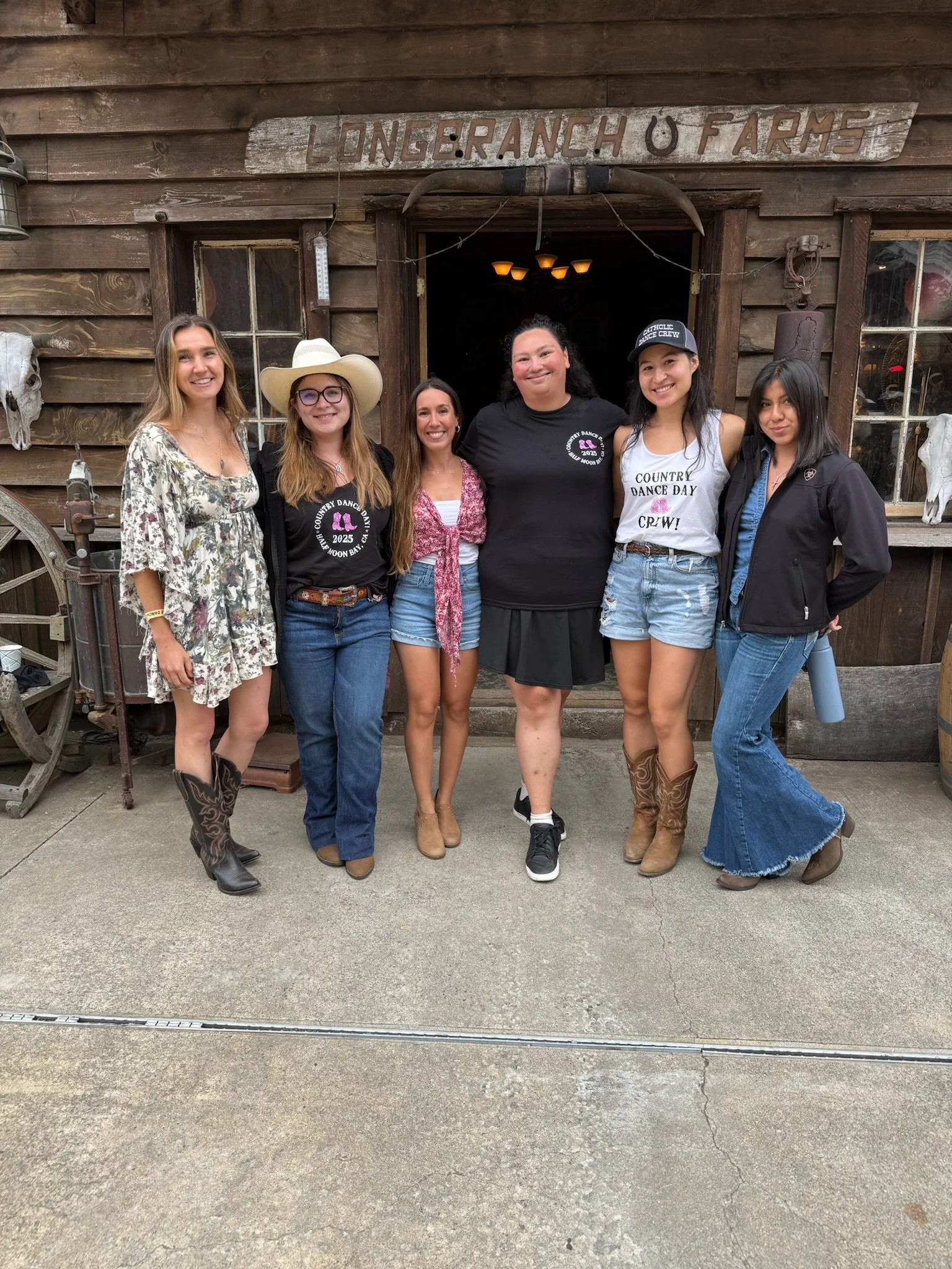 Six women standing in front of a rustic wooden barn with a sign that reads 'Long Ranch U Farms.' They are dressed in casual and western-style clothing, with some wearing cowboy boots, hats, and denim. The women are smiling and posing for the photo.