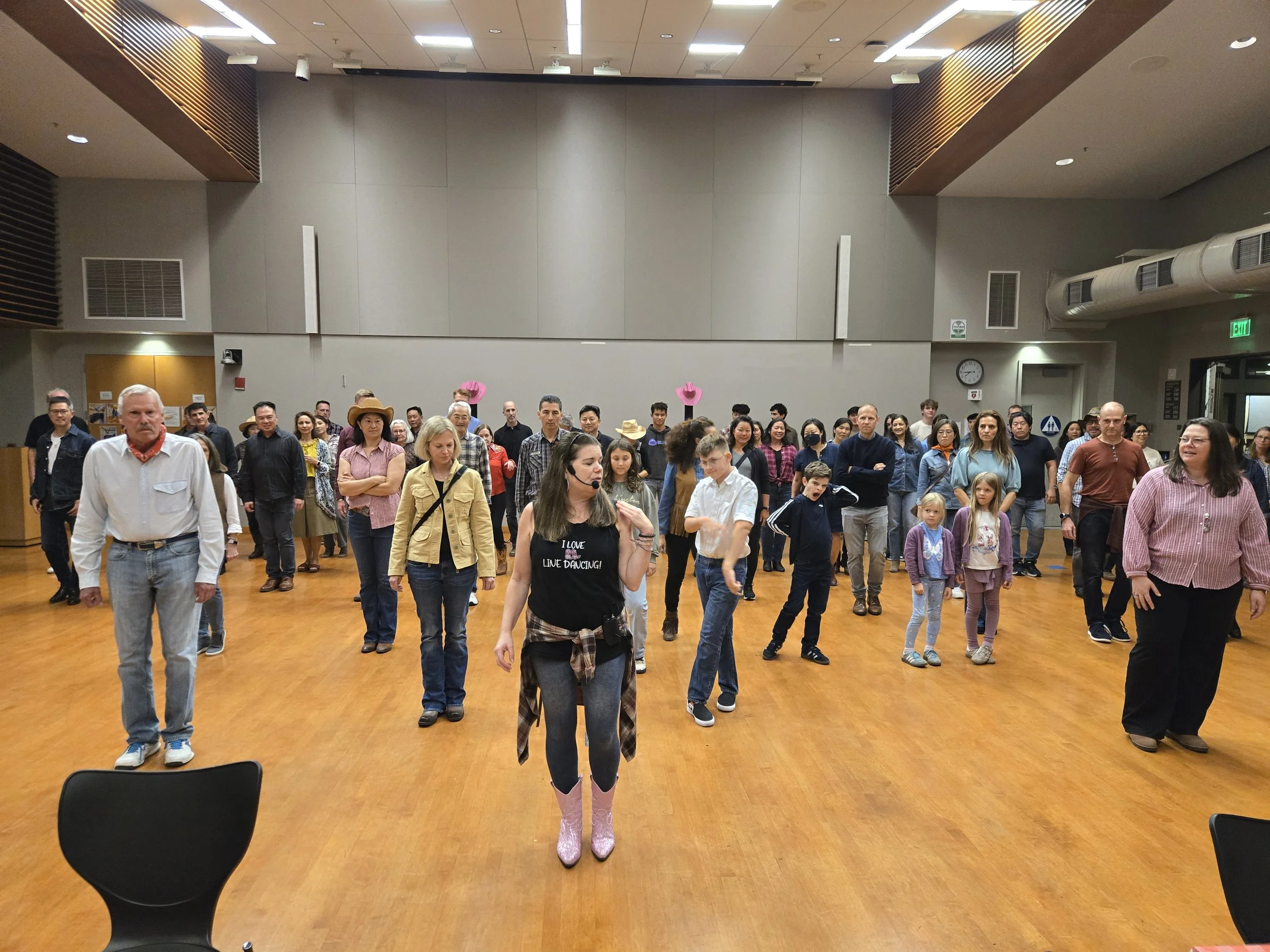 A diverse group of people is participating in a line dancing event in a spacious indoor hall with a wooden floor and high ceiling.