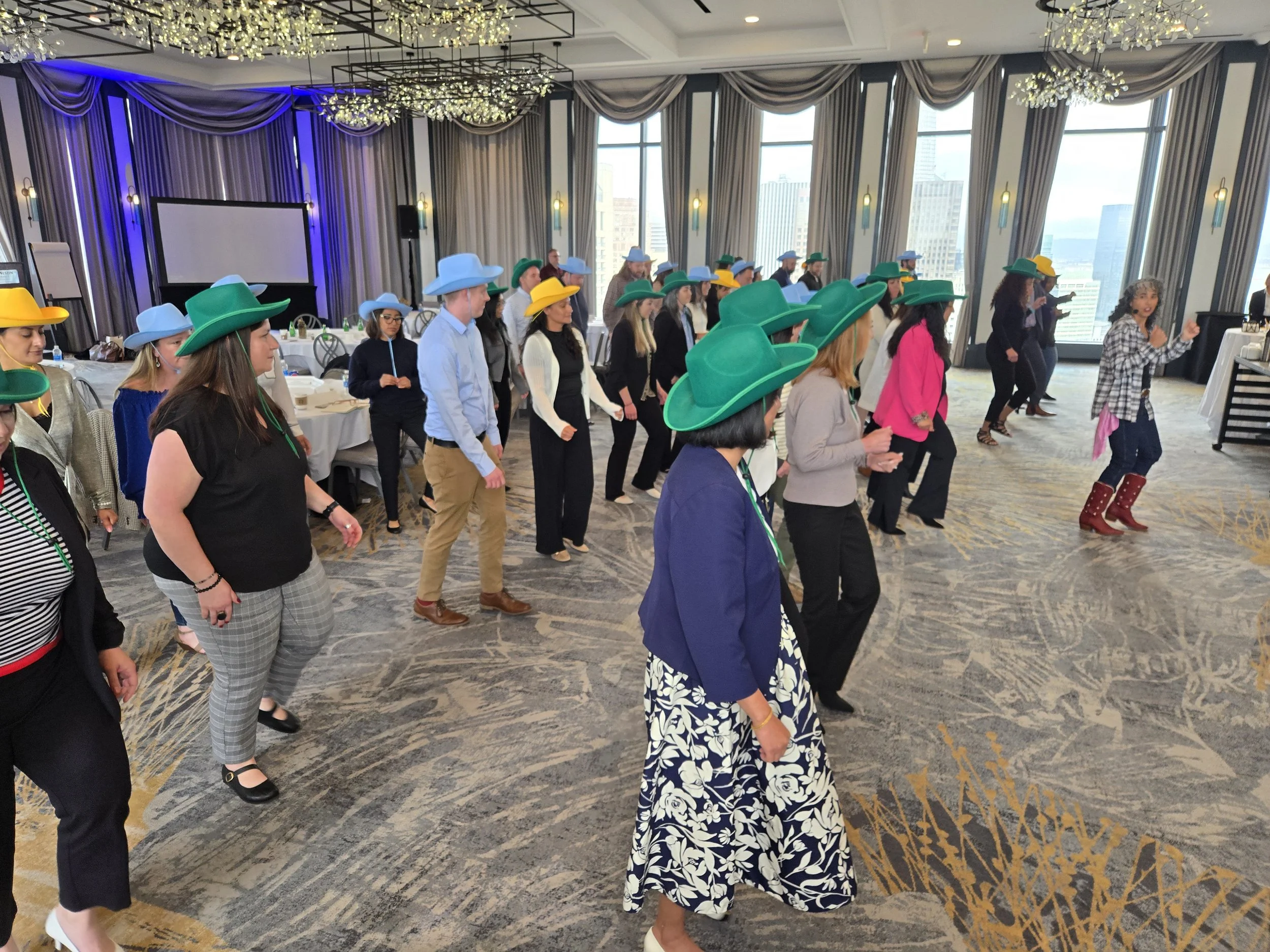 A corporate party at a hotel in San Francisco with DJ Shivers and Line Dancing Lisa showing a group of people dancing in a conference room decorated with chandeliers and large windows, many wearing cowboy hats and colorful clothing.