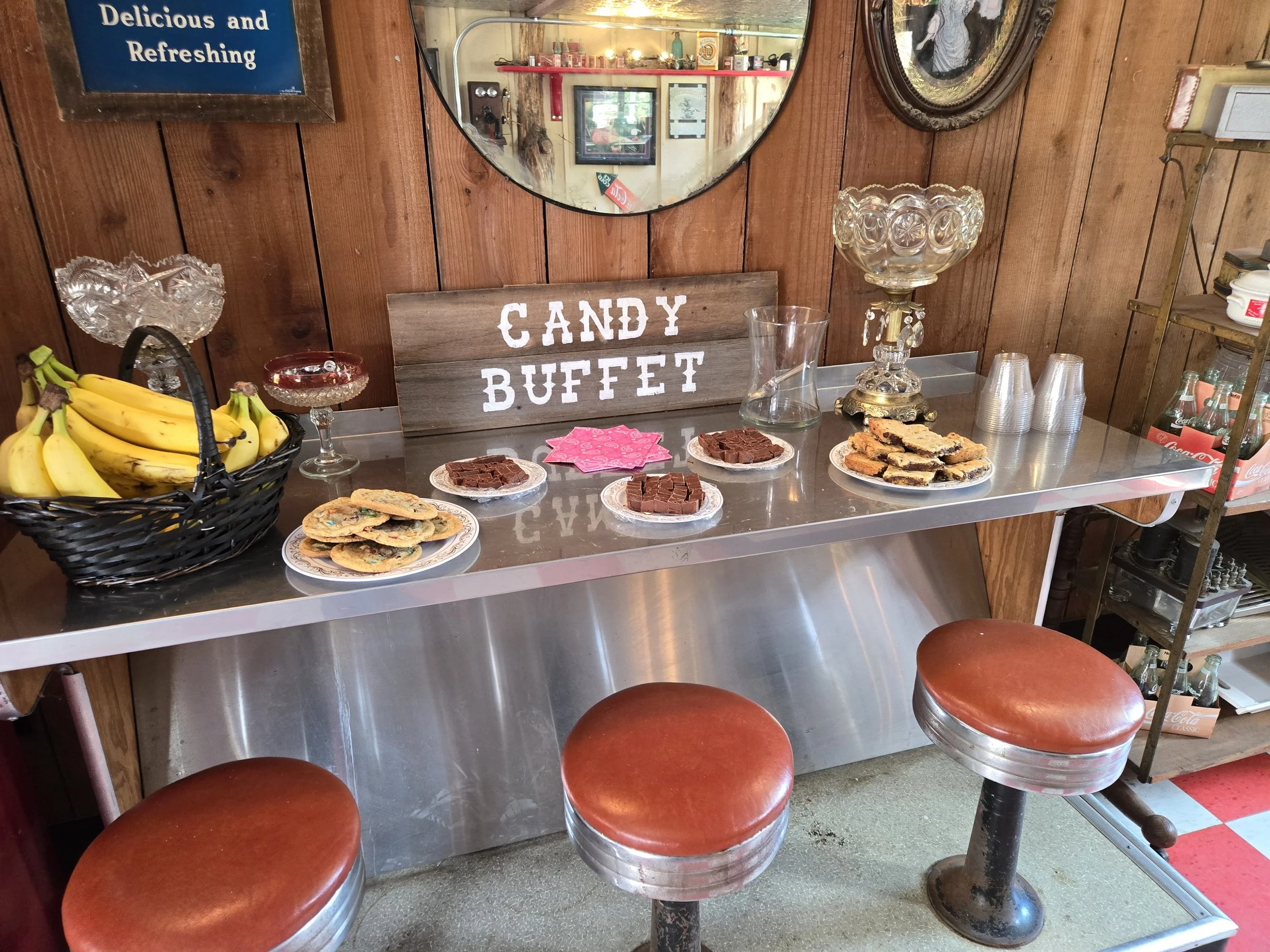 A candy buffet table with various treats including chocolate chip cookies, brownies, and cookie bars. There are bananas in a basket and a sign that reads 'CANDY BUFFET.' The table is decorated with glass candy dishes, a pitcher, and a large decorative glass vase. Behind the table is a wood-paneled wall with a mirror and a framed sign that says 'Delicious and Refreshing.'