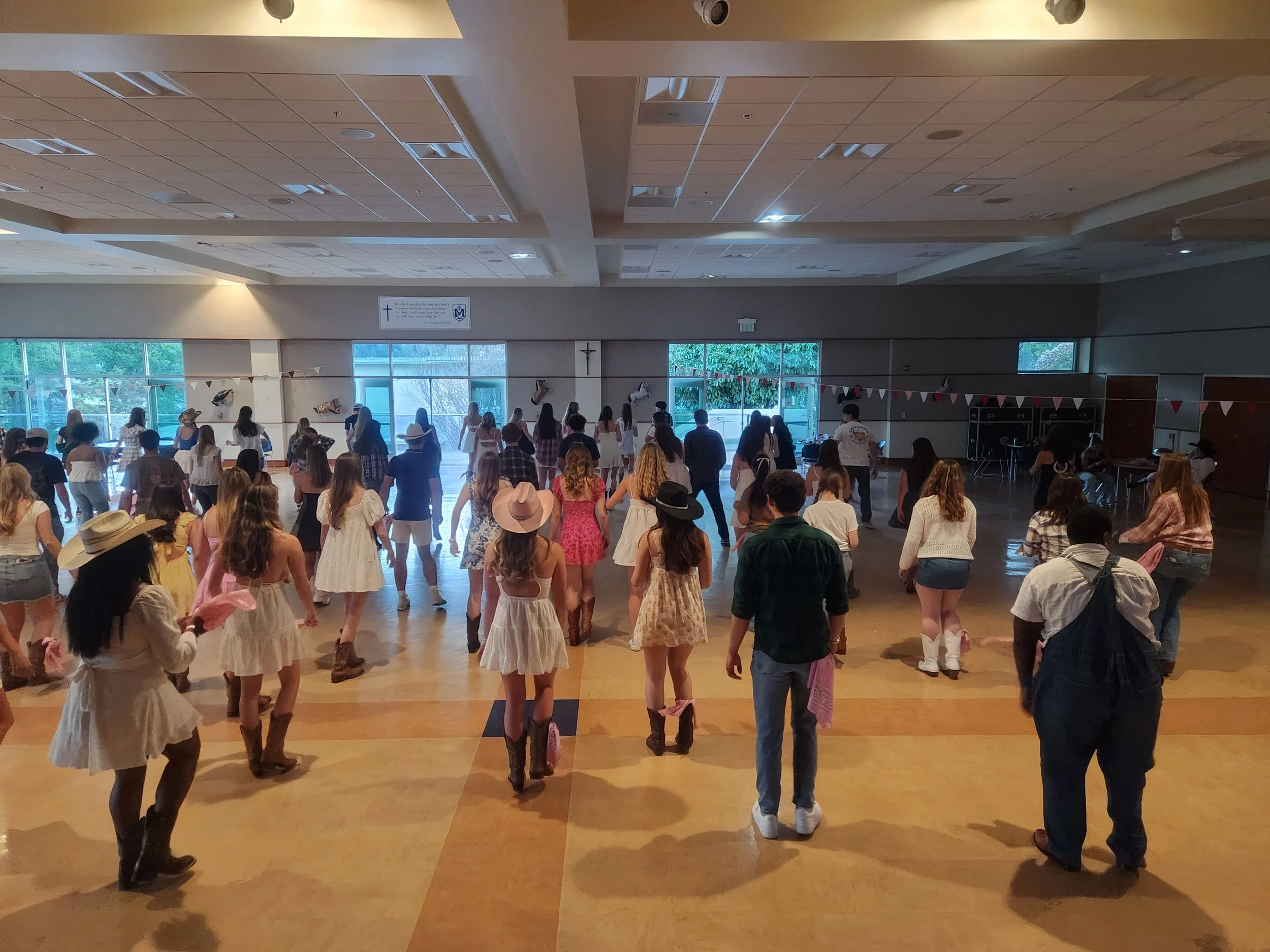 A private school dance event by DJ Shivers. Country-themed line dance with women in dresses and cowboy hats, men in casual wear, in a decorated indoor hall with bunting and large windows.