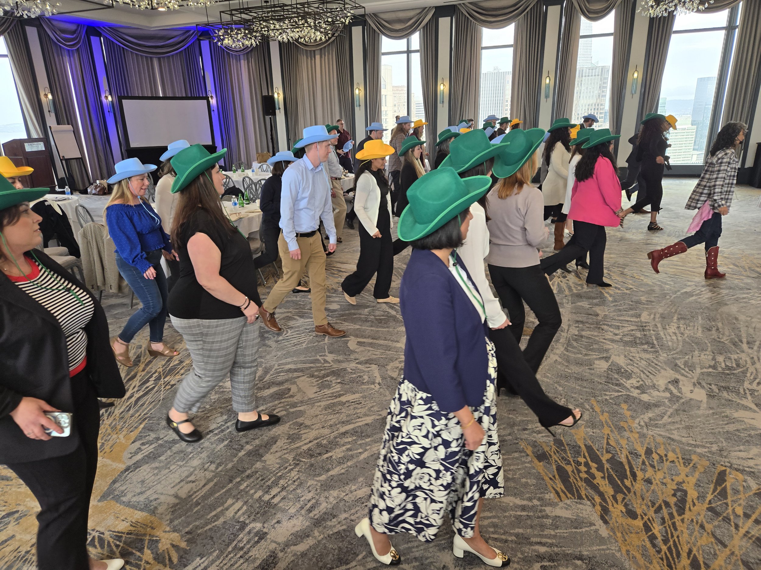 DJ Shivers and Line Dancing Lisa private Corporate Event. People dancing in a room with large windows and city view, wearing cowboy hats in various colors, at a social or corporate event.