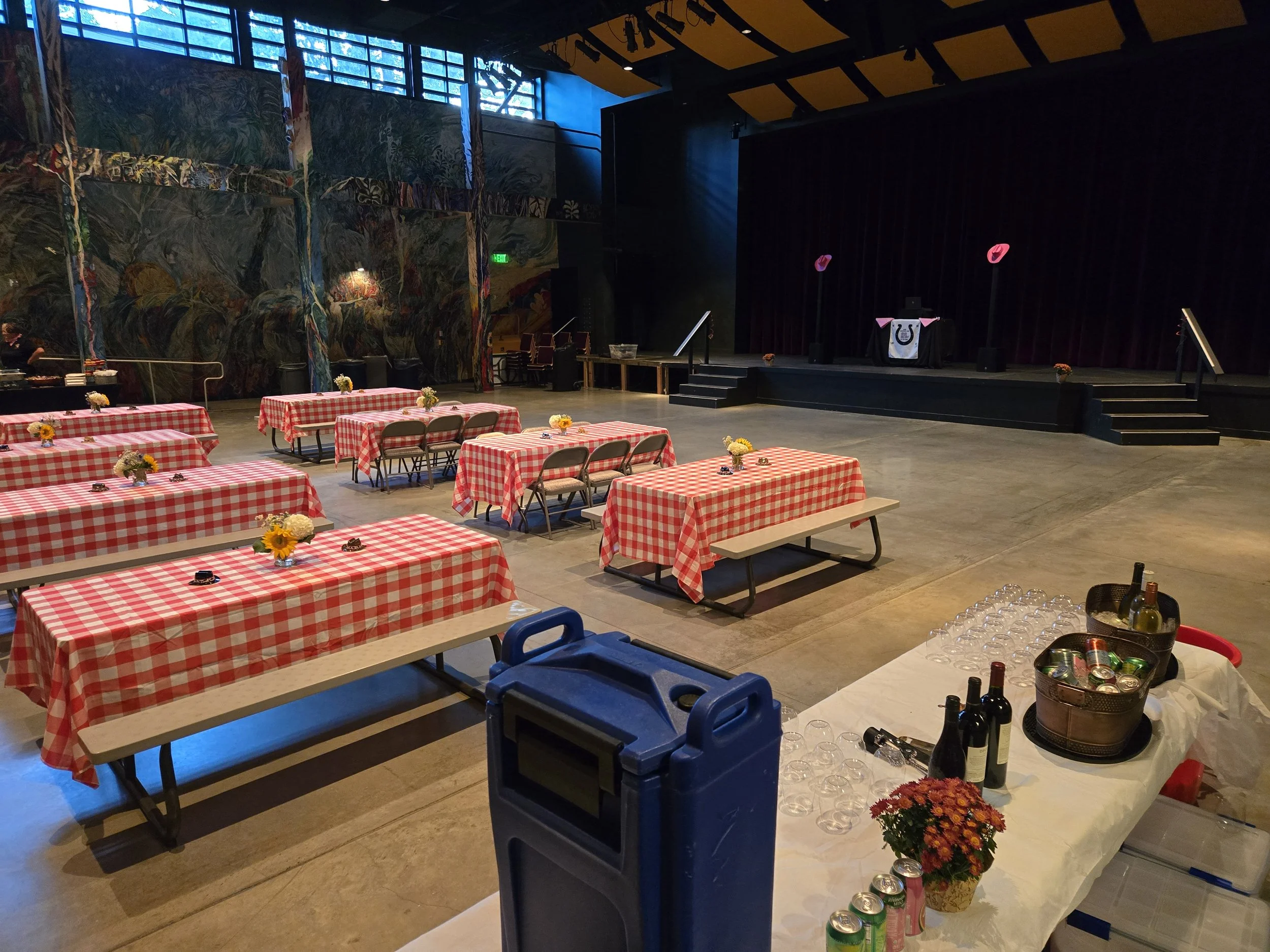 An indoor event space with tables covered in red and white checkered tablecloths, decorated with small flower bouquets, set up for a gathering or party. There is a stage with a DJ booth and decorative balloons, and a table with bottles of wine, cans, and glasses in the foreground.