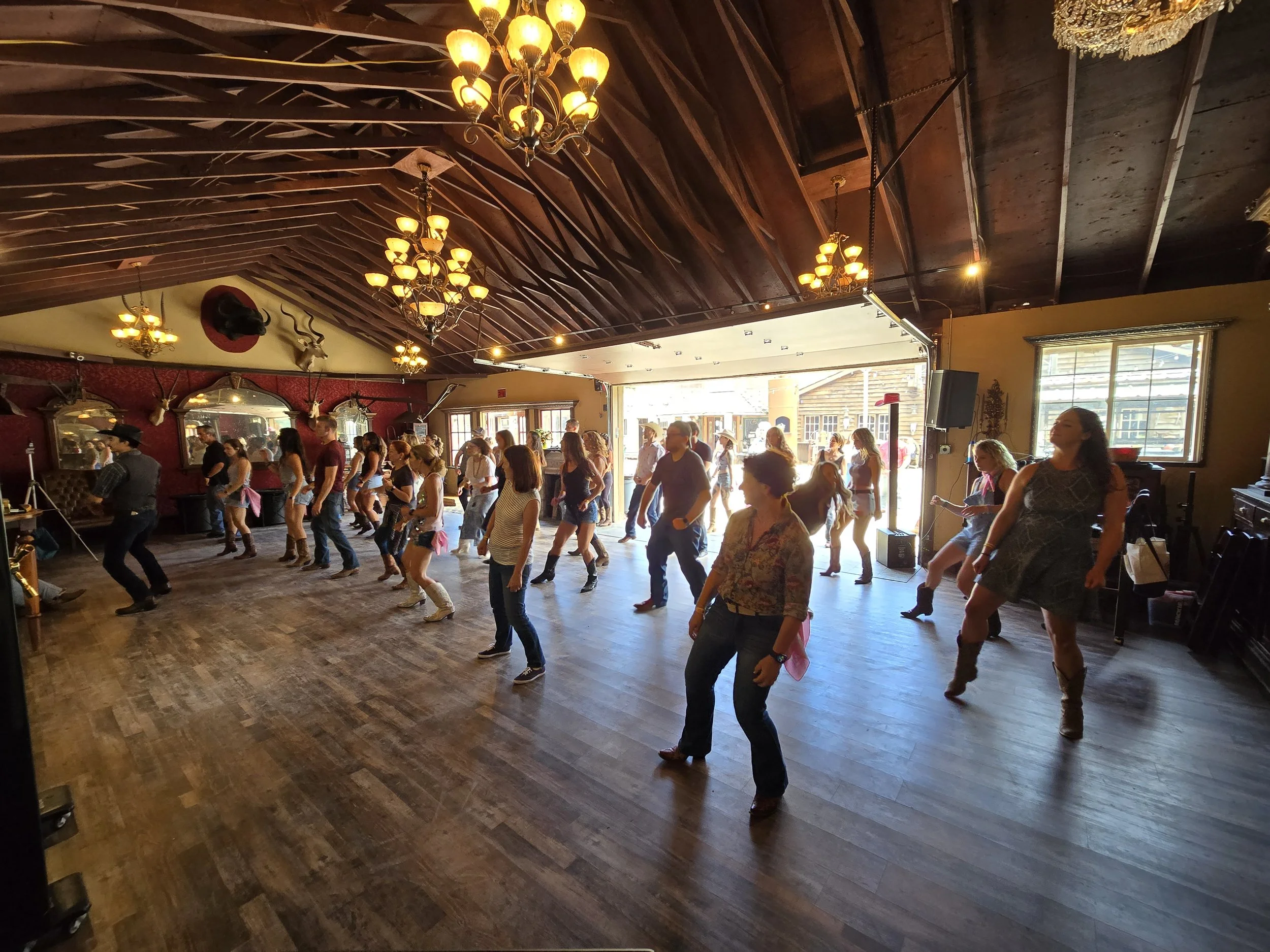 Group of people line dancing inside a rustic venue with wood floors, chandeliers, and mounted animal heads, with sunlight streaming in from the outside at Country Dance Day In Half Moon Bay. Featuring Line Dancing Lisa and DJ Shivers.