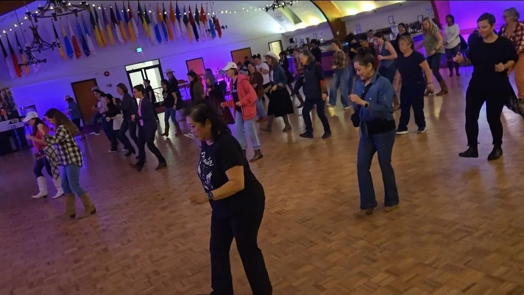 DJ Shivers and Line Dancing Lisa at the United Irish Cultural Center 1 Friday per month in San Francisco. Group of people line dancing in a large hall with a wooden floor, colorful lighting, and flags hanging from the ceiling.
