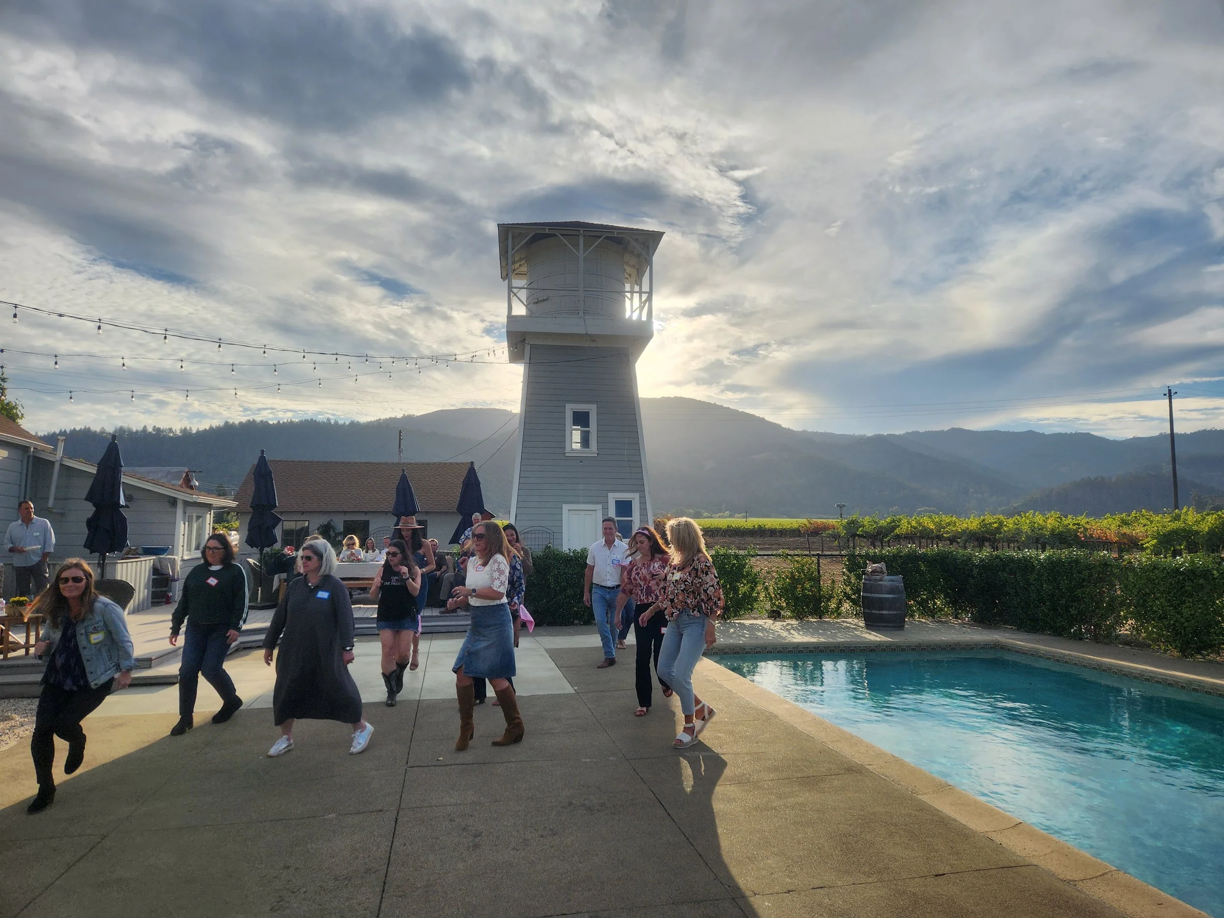 Winery in Napa private event for members. Line Dancing Lisa instructing people line dancing on a patio near a pool with a water tower in the background, under a partly cloudy sky with mountains in the distance.