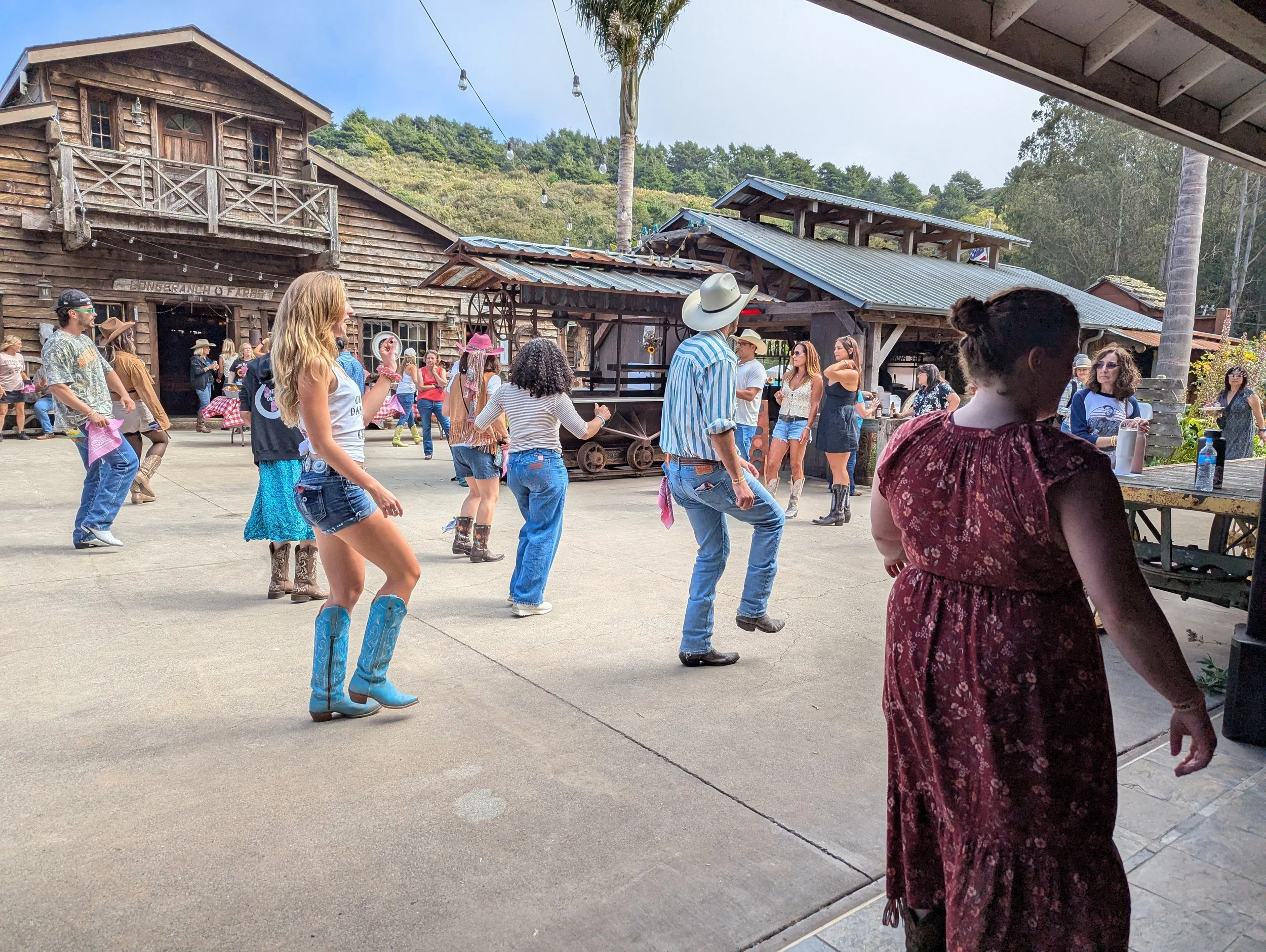 People enjoying a line dance outdoors in a rustic setting with wooden buildings, some dressed in country or Western attire, with mountains and trees in the background. Country Dance Day in Half Moon Bay Event. Happens every August at Long Branch