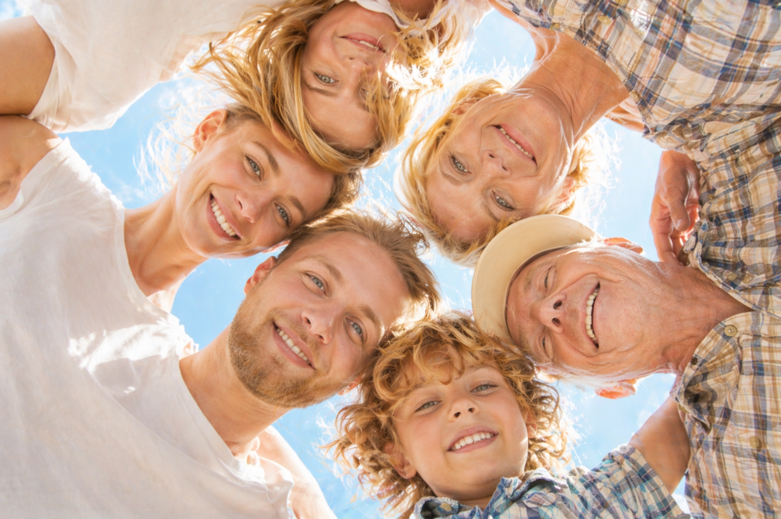 A group of six smiling people, including children, gathered in a circle outdoors, looking down at the camera with blue sky and sunlight above.