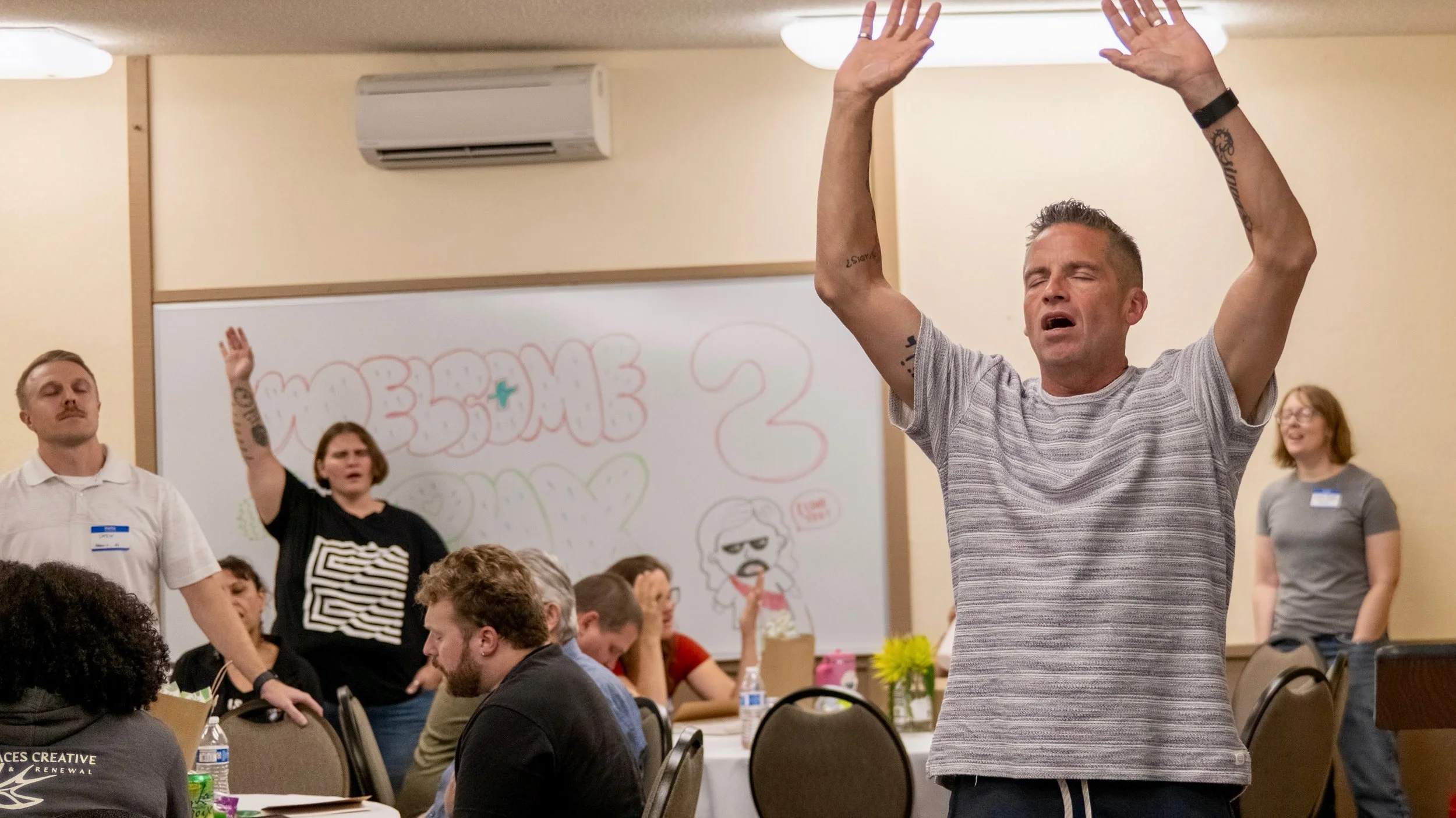 A man with closed eyes and raised arms appears to be singing or praying at an indoor gathering. Other people are sitting at tables and standing in the background, some with eyes closed or looking around. A whiteboard with colorful writing and drawings is visible behind them.