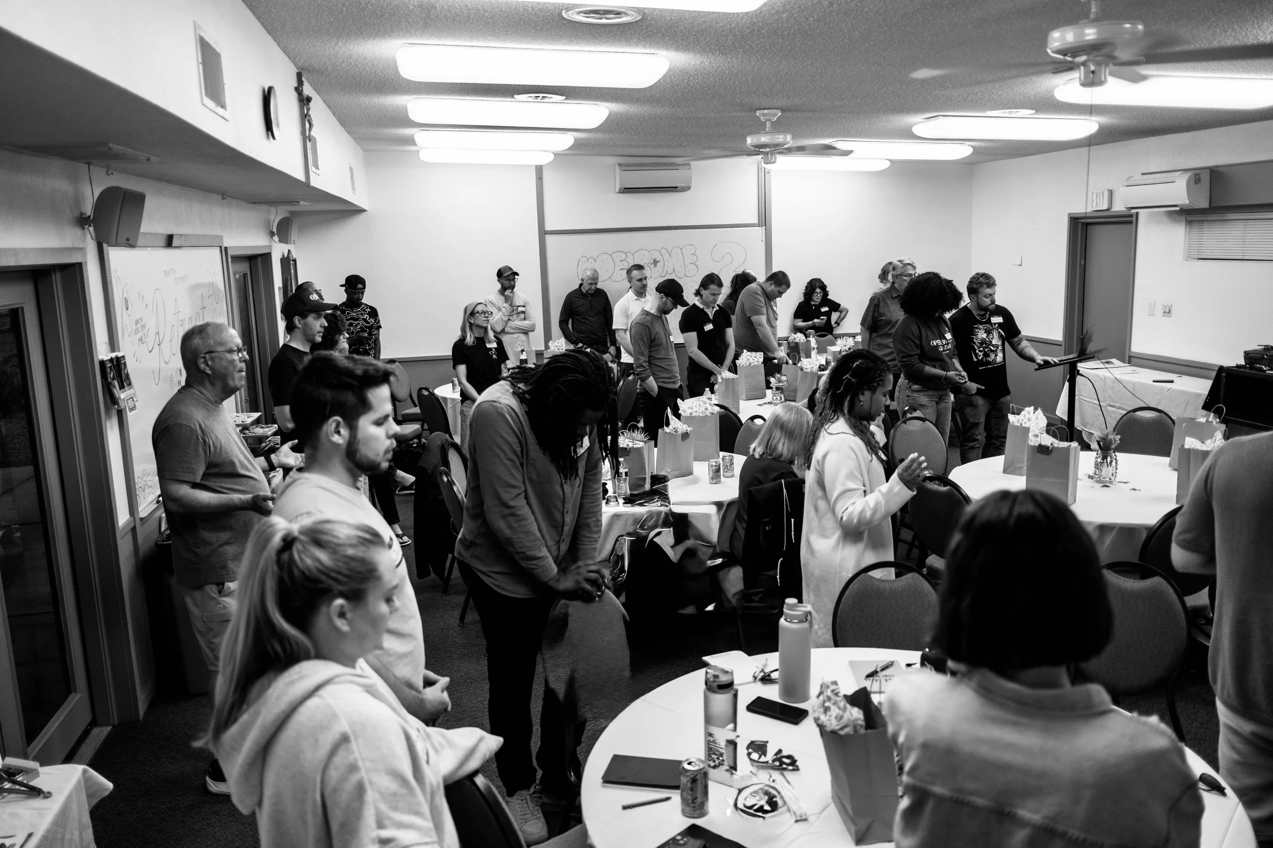 People gathered in a room with tables, some of which are adorned with gift bags and flowers, during a memorial or prayer session; individuals are standing with eyes closed or heads bowed, in a moment of reflection.